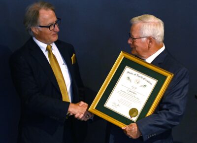 Two men in suits shake hands. The main on the right holds a gold-framed certificate.