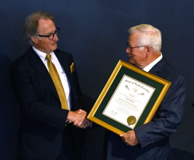 Two men in suits shake hands. The main on the right holds a gold-framed certificate.