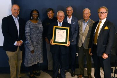 A group of people stand in a row. The man in the center holds a gold-framed certificate.