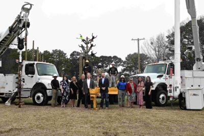 A group of people standing outdoors between two white utility trucks with bucket lifts, near a newly planted tree.