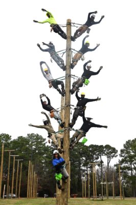 Electric lineworkers posed climbing a tall utility pole in coordinated positions, wearing safety gear and harnesses, outdoors near a training field.