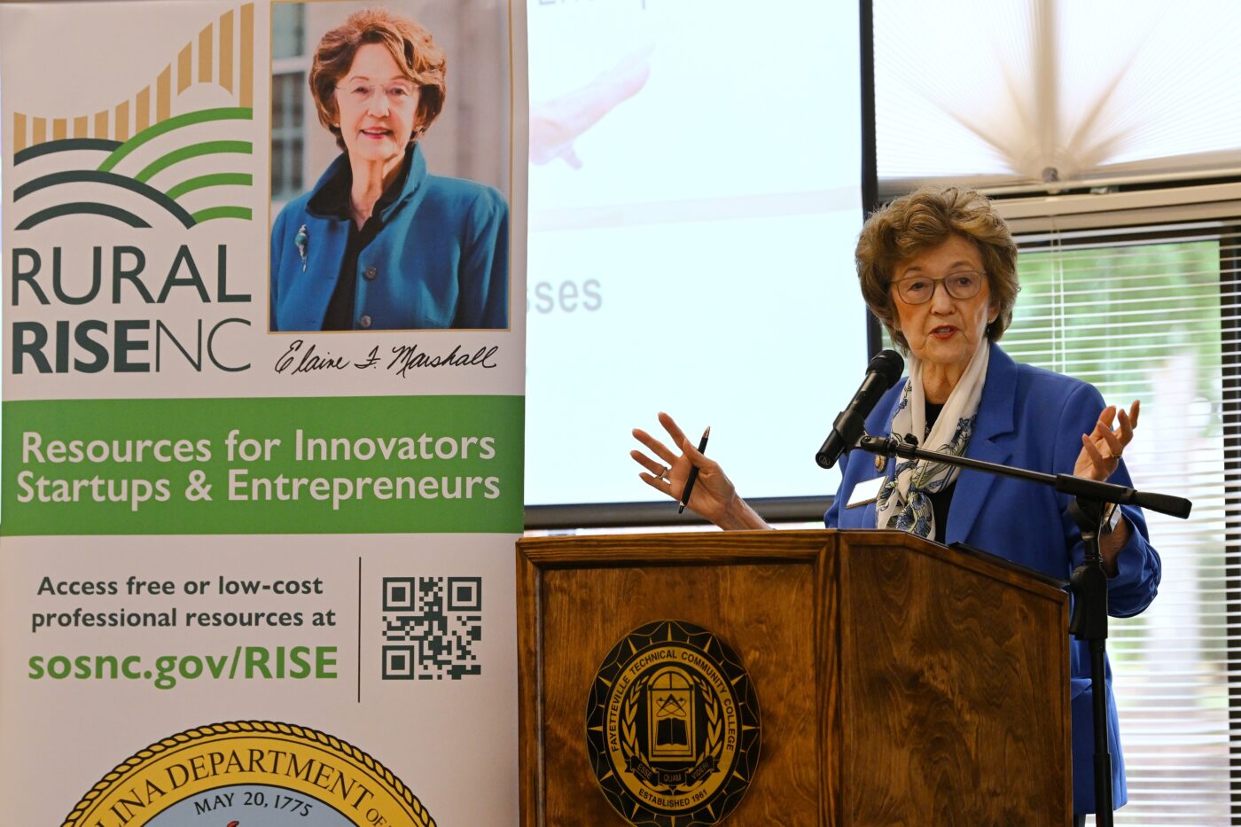 NC Secretary of State Elaine Marshall speaks and stands at a podium. A banner behind her promotes the NC Rural RISE small business program.