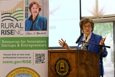 NC Secretary of State Elaine Marshall speaks and stands at a podium. A banner behind her promotes the NC Rural RISE small business program.