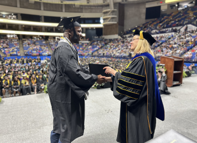 A woman in doctoral robes hands a degree folder to and shakes hands with a graduate in a cap and gown.