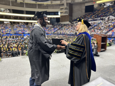 A woman in doctoral robes hands a degree folder to and shakes hands with a graduate in a cap and gown.