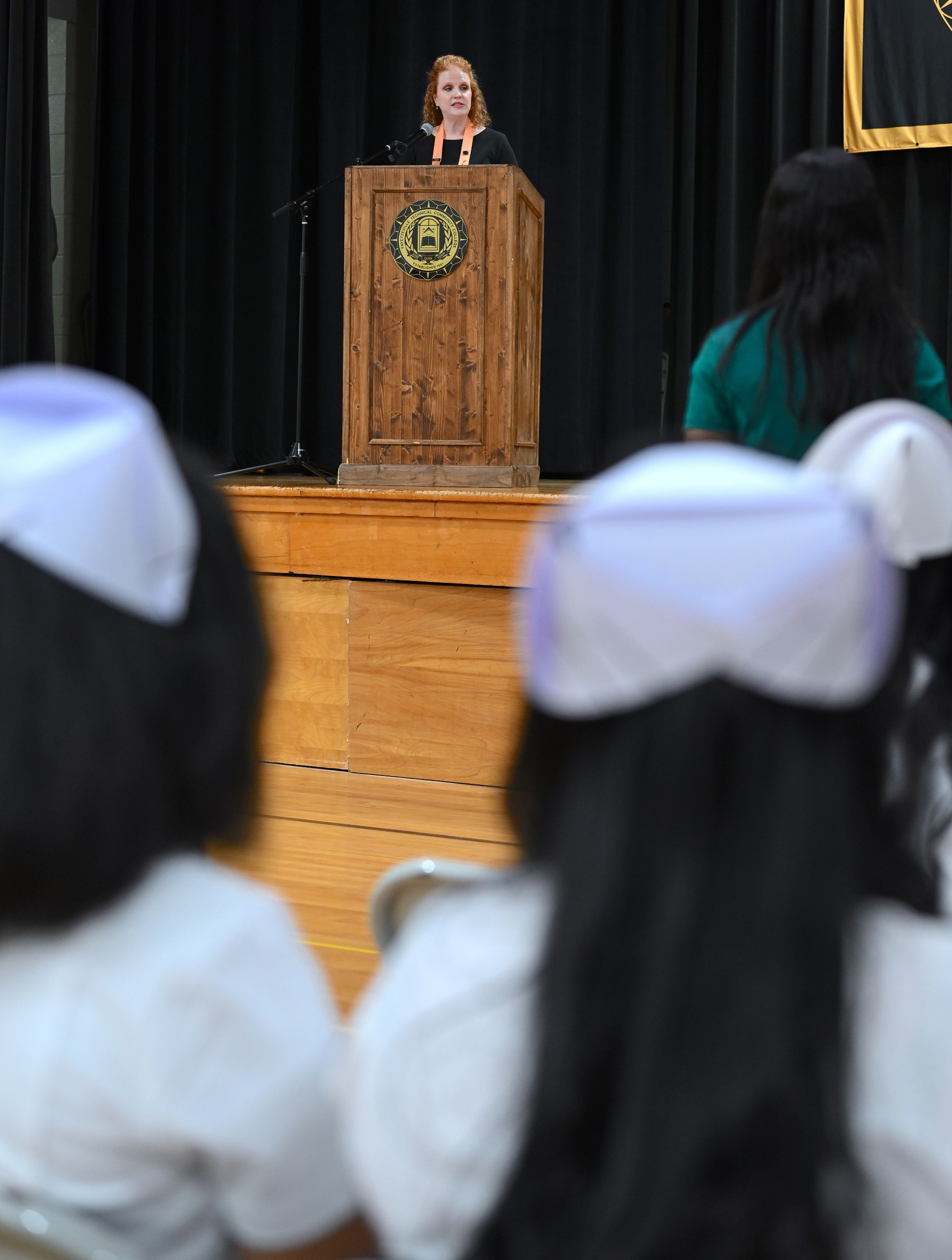 Melanie Stephens speaks at the podium on stage. Graduates in white Nursing caps and uniforms are seen from behind in the foreground.
