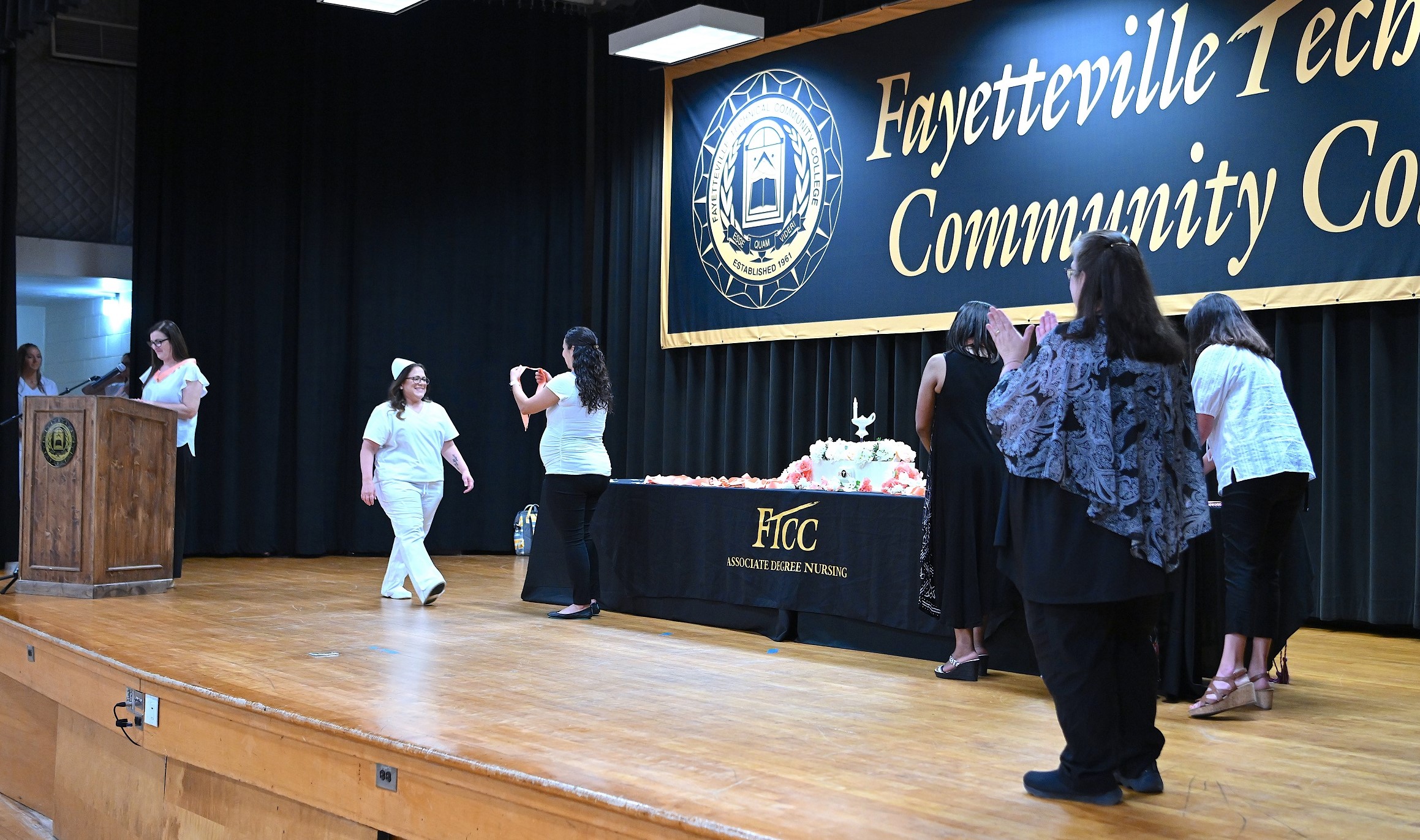 A wide photo of the stage as a graduate crosses to receive her pin from instructors.