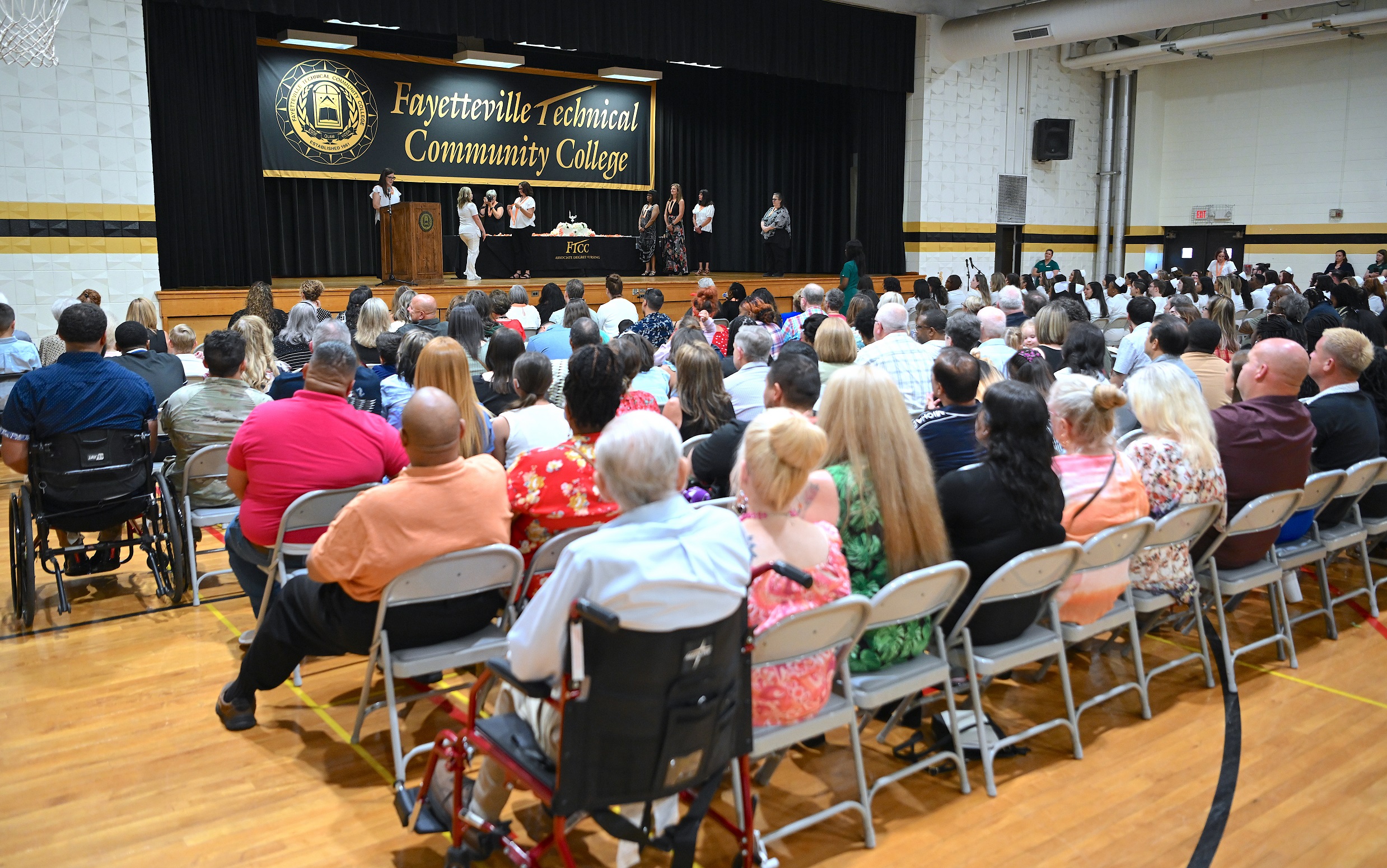 A photo taken from behind the audience shows the stage as a graduate walks to receive her pin in the background.