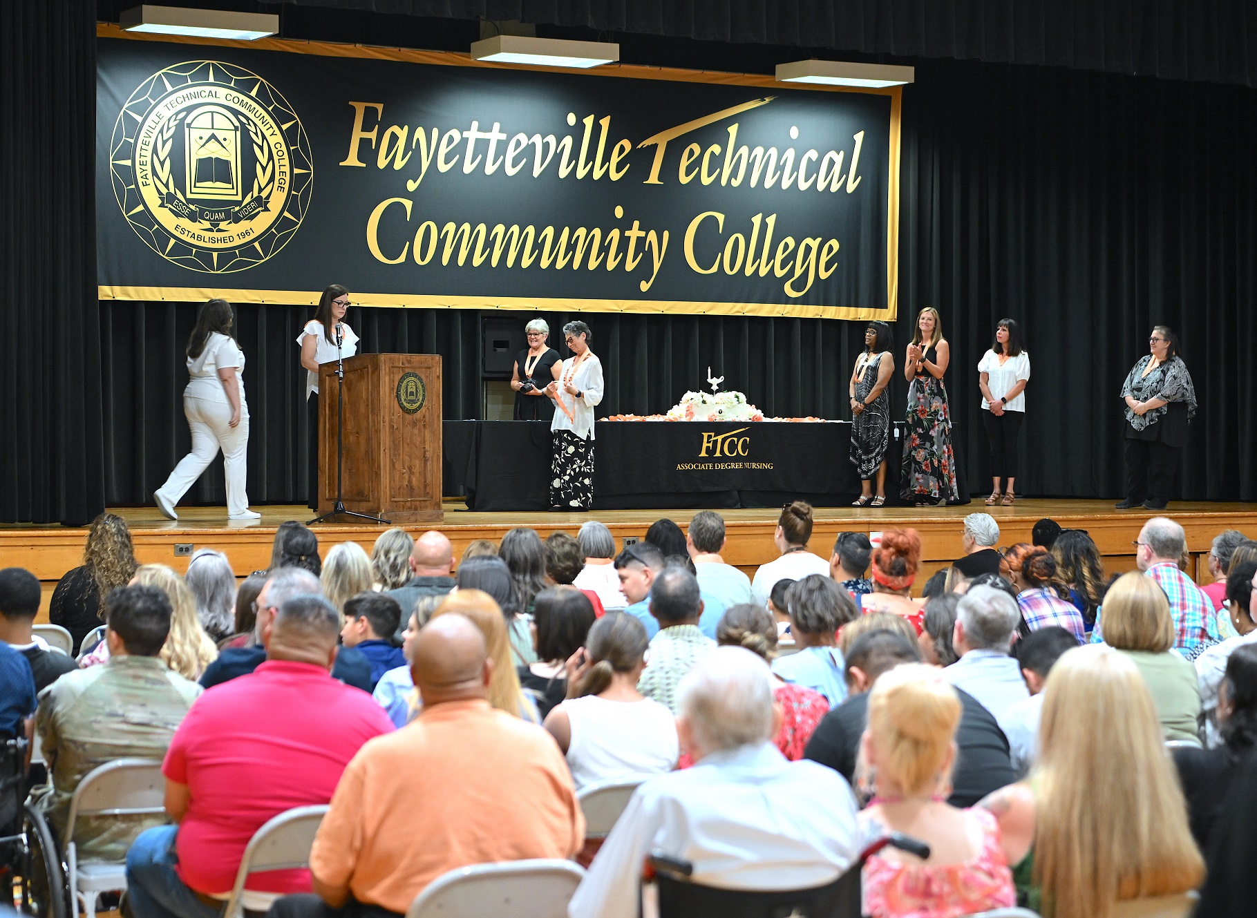 A photo taken from behind the audience shows the stage as a graduate walks to receive her pin in the background.