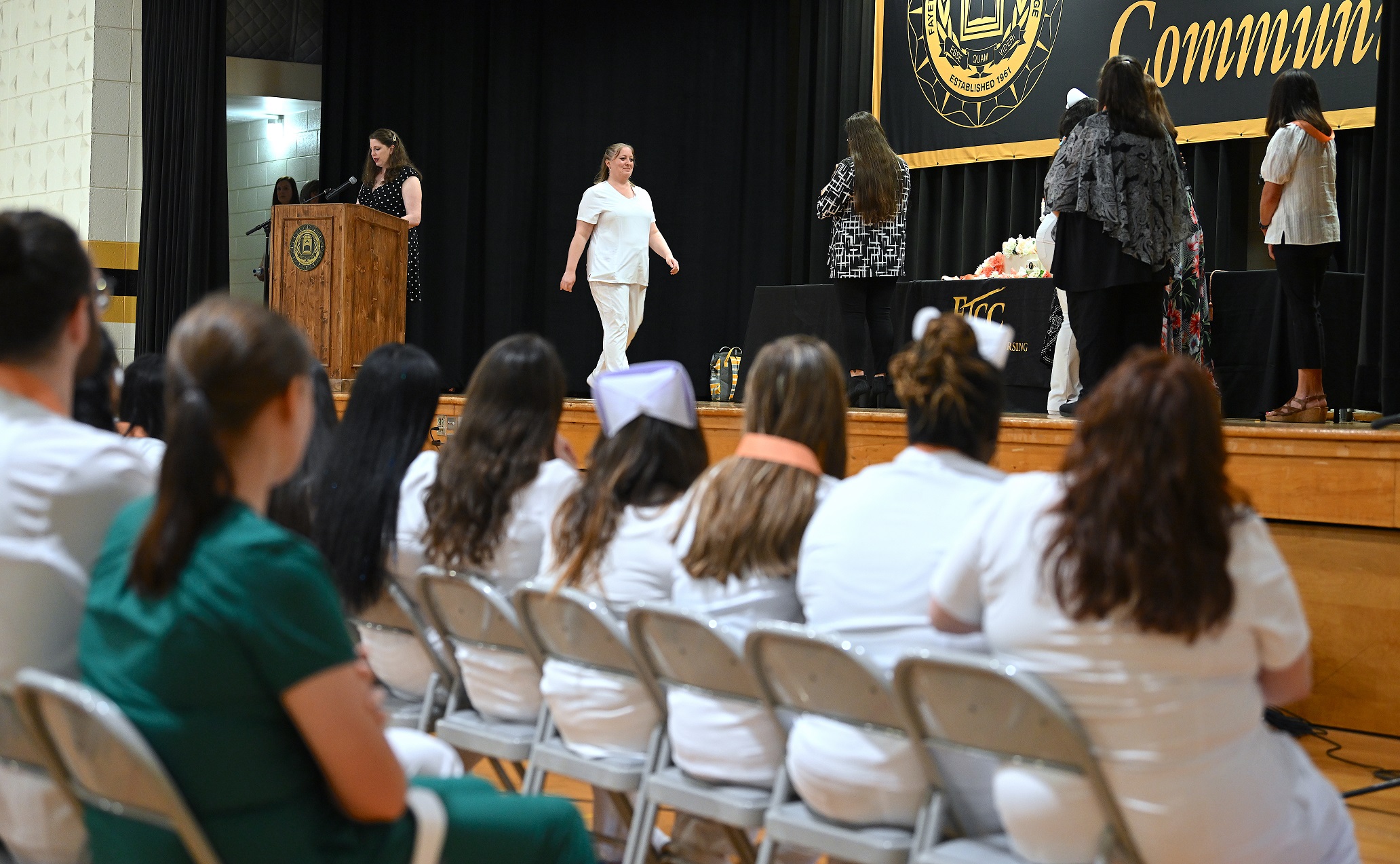 A graduate crosses the stage to receive her pin. Rows of graduates seated in front of the stage are visible in the foreground.