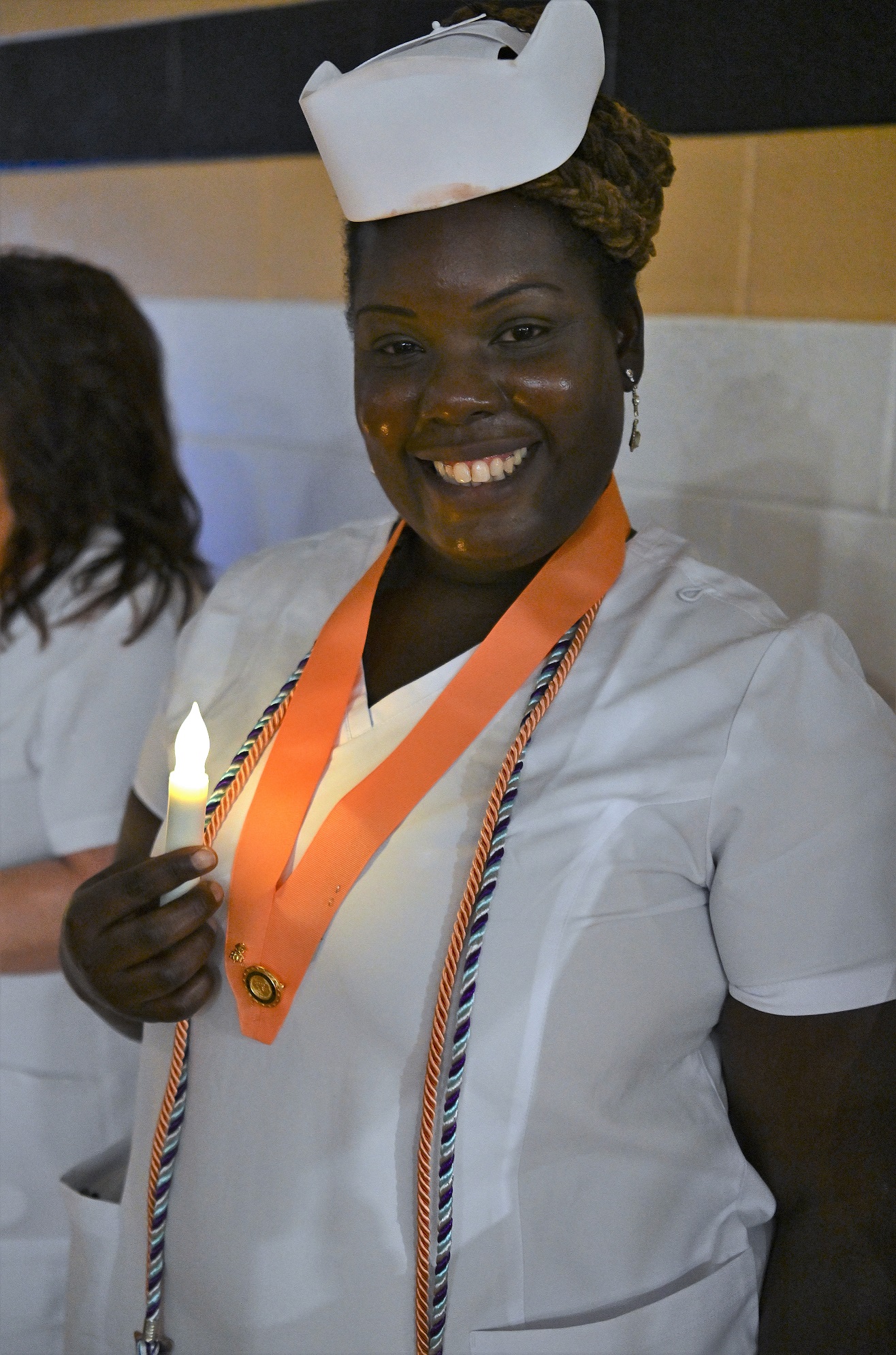 A close-up of a Nursing graduate, wearing a peach-colored pin ribbon and two cords. She is holding a battery-operated candle.