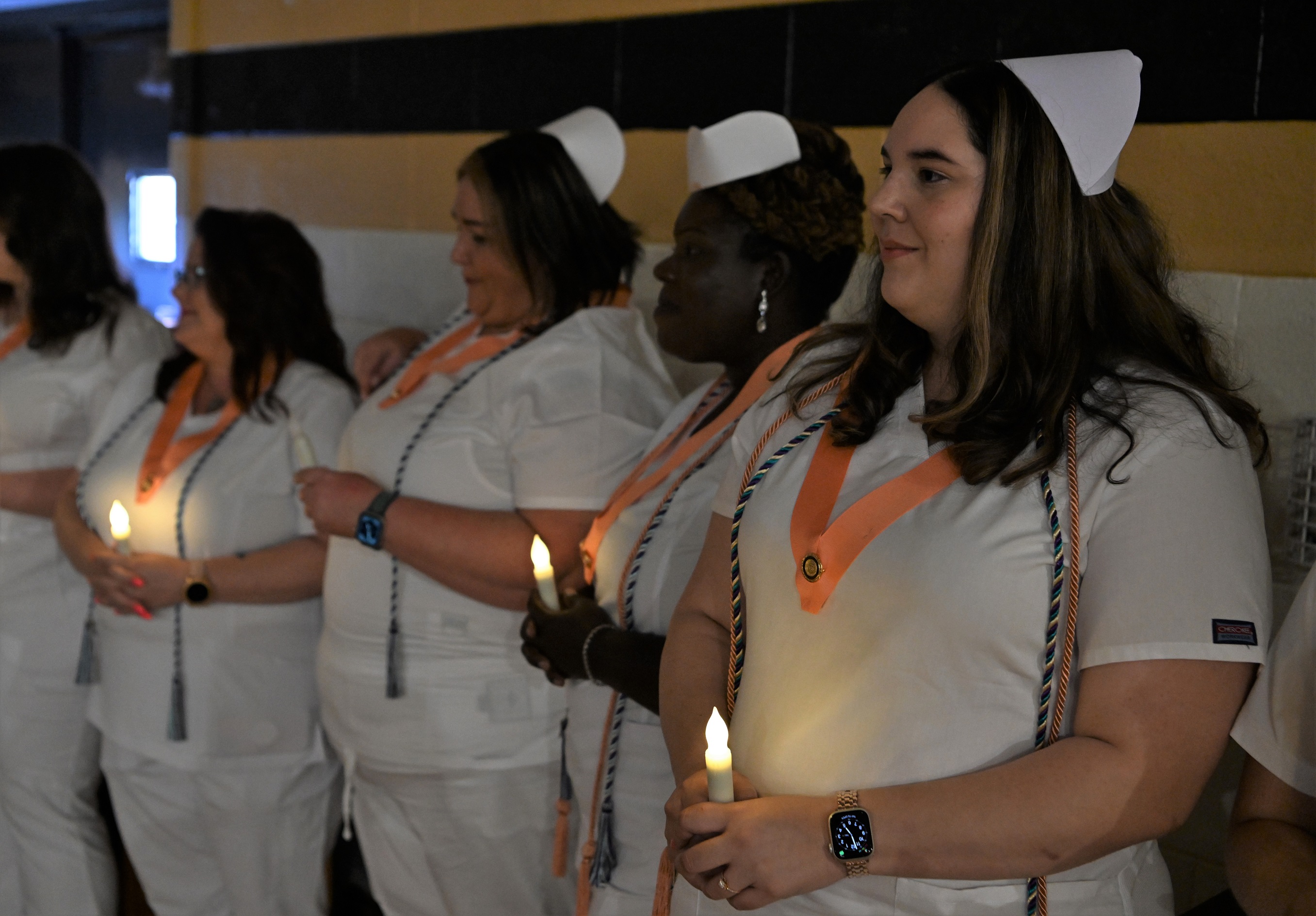 Graduates stand against the wall. They are each holding a battery-operated candle and are wearing peach-colored pin ribbons.