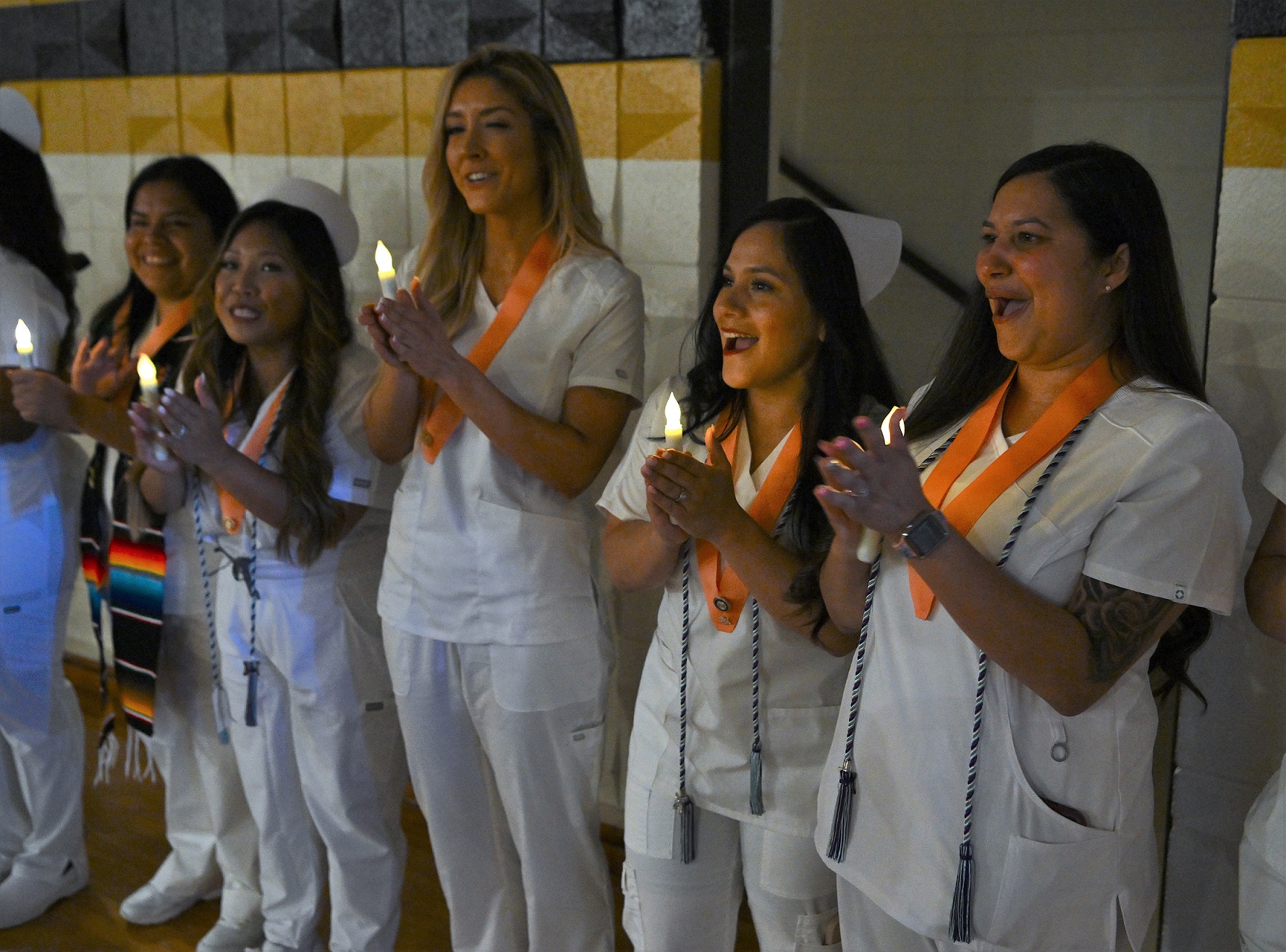Graduates stand against the wall. They are each holding a battery-operated candle and are wearing peach-colored pin ribbons.