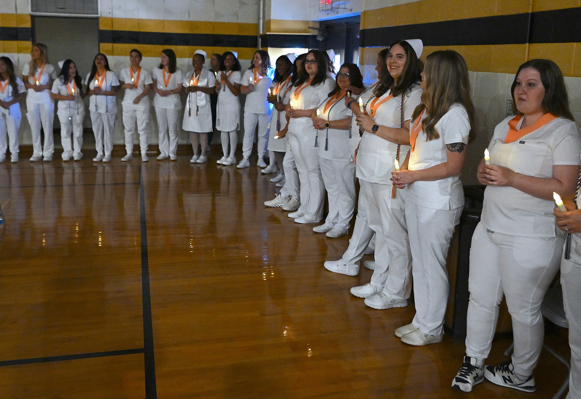 Graduates stand against the wall. They are each holding a battery-operated candle and are wearing peach-colored pin ribbons.