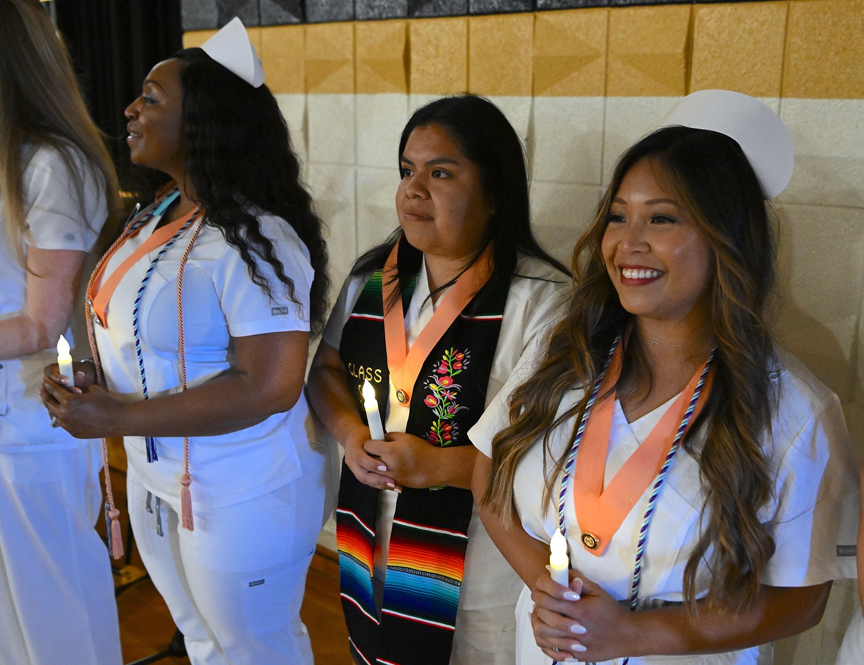 Graduates stand against the wall. They are each holding a battery-operated candle and are wearing peach-colored pin ribbons.