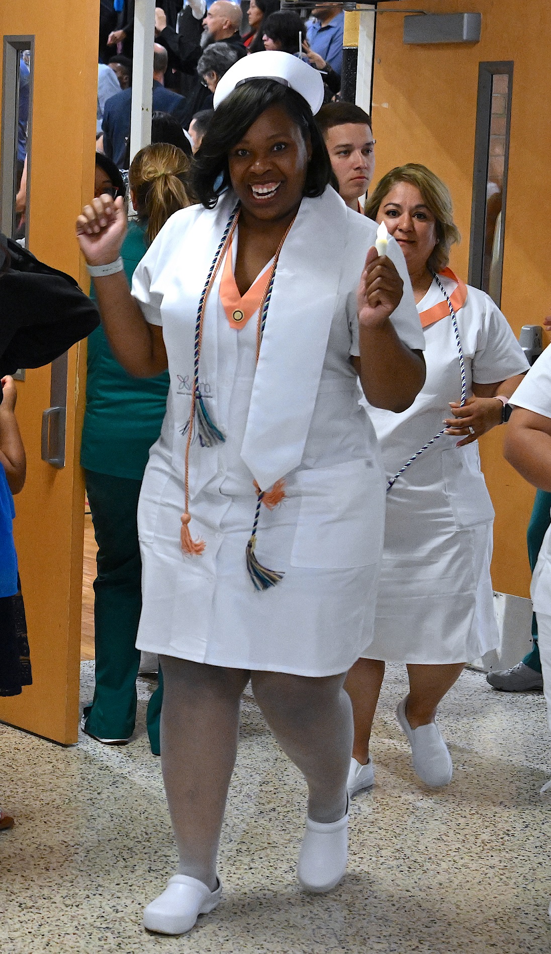 A graduate cheers as she walks out of the gymnasium. She is wearing a white stole, two cords and a peach-colored pin ribbon over her white nurse uniform.