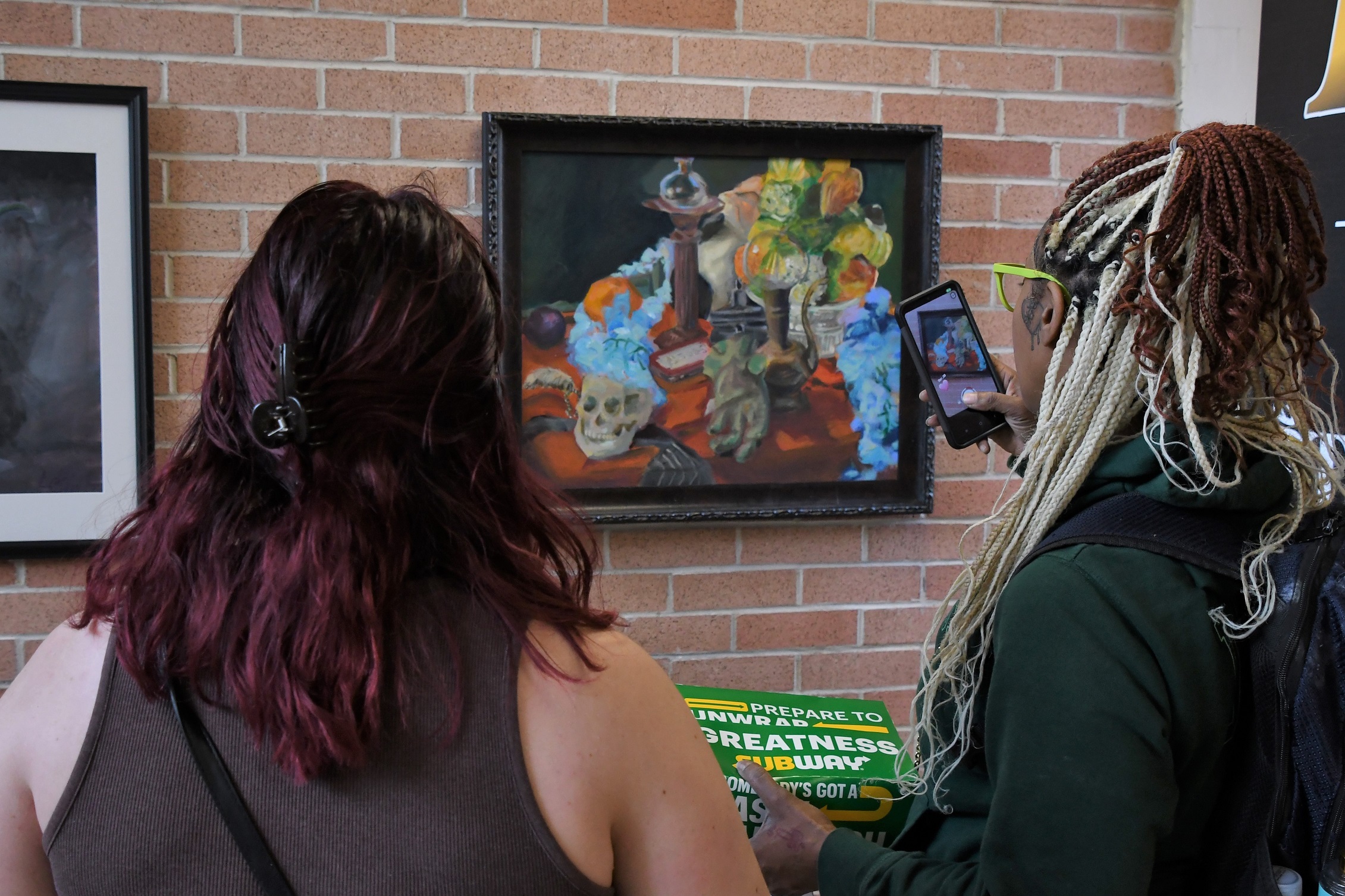 Two students look at a paining. The student on the right is taking a photo of the painting with a cell phone.