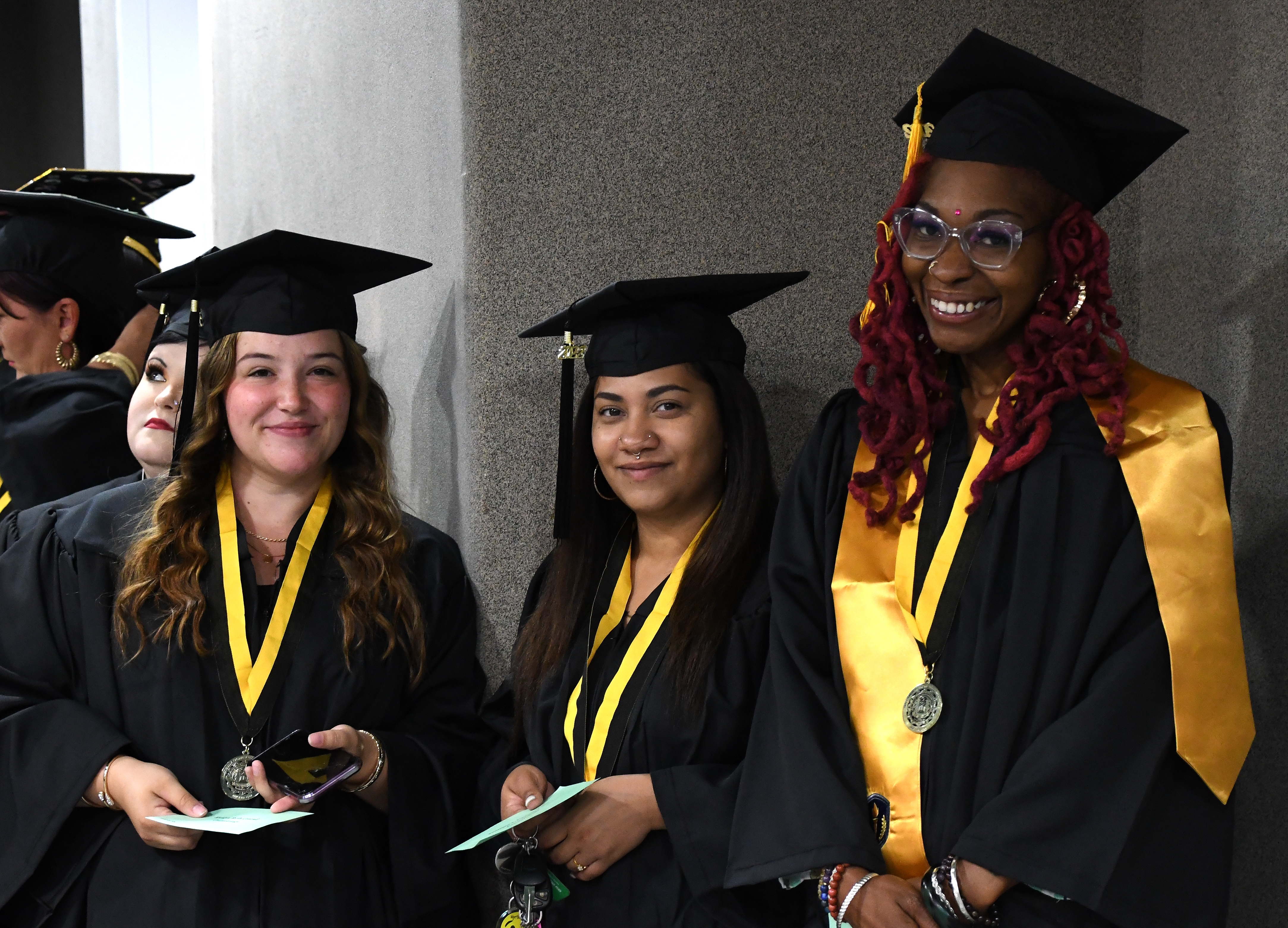Three graduates smile at the camera while they wait in the hallway before walking in the graduation processional.