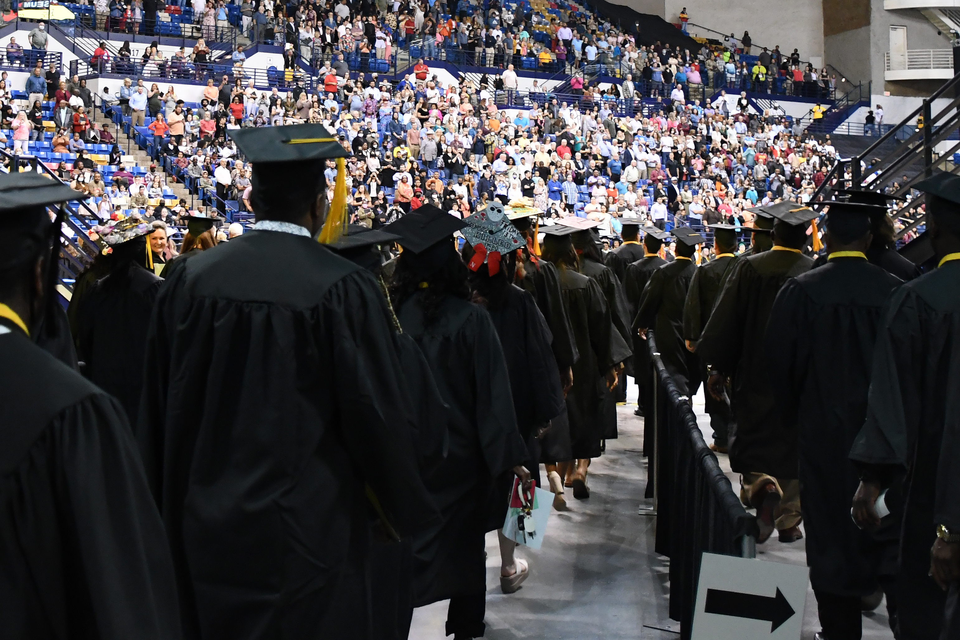A photo taken inside the tunnel leading to the arena floor at graduation. The backs of several graduates is visible in the foreground, and the crowd is visible in the background.