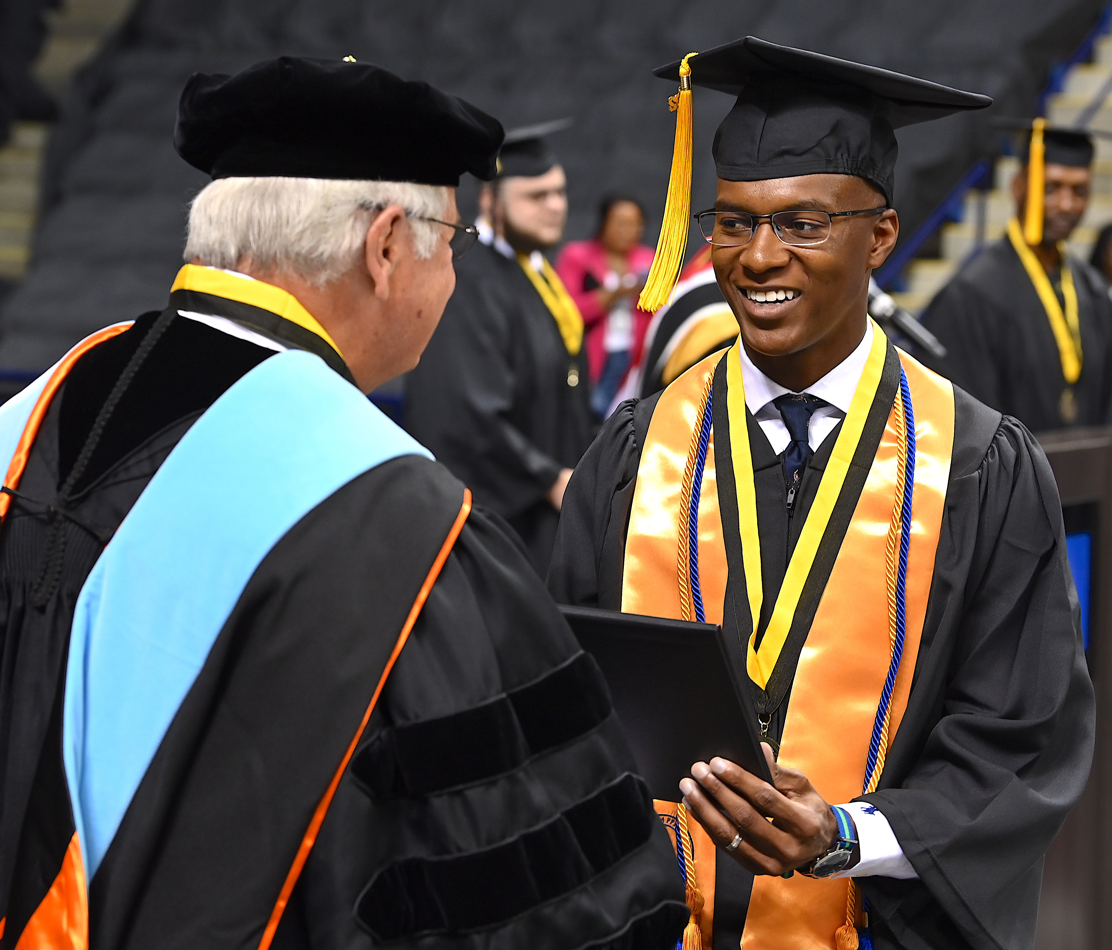 A smiling graduate wearing glasses, a gold stole, a gold cord and blue cord and a medal on a black and gold ribbon around his neck, accepts his degree from Dr. Keen.