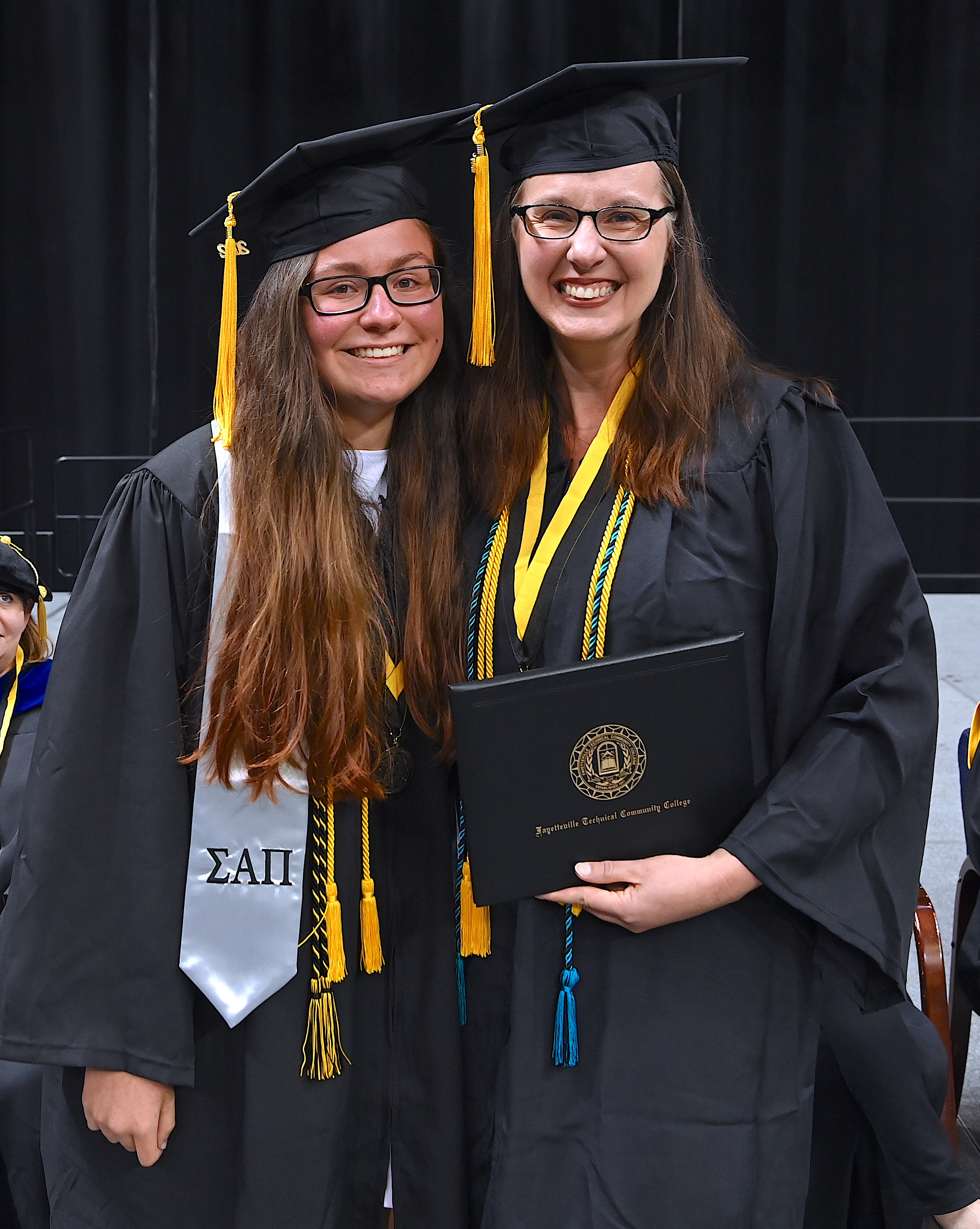 Jenna Warnock and her mother, Jennifer Warnock pose side-by-side. They are both dressed in their caps and gowns, and Jennifer is holding a degree portfolio.