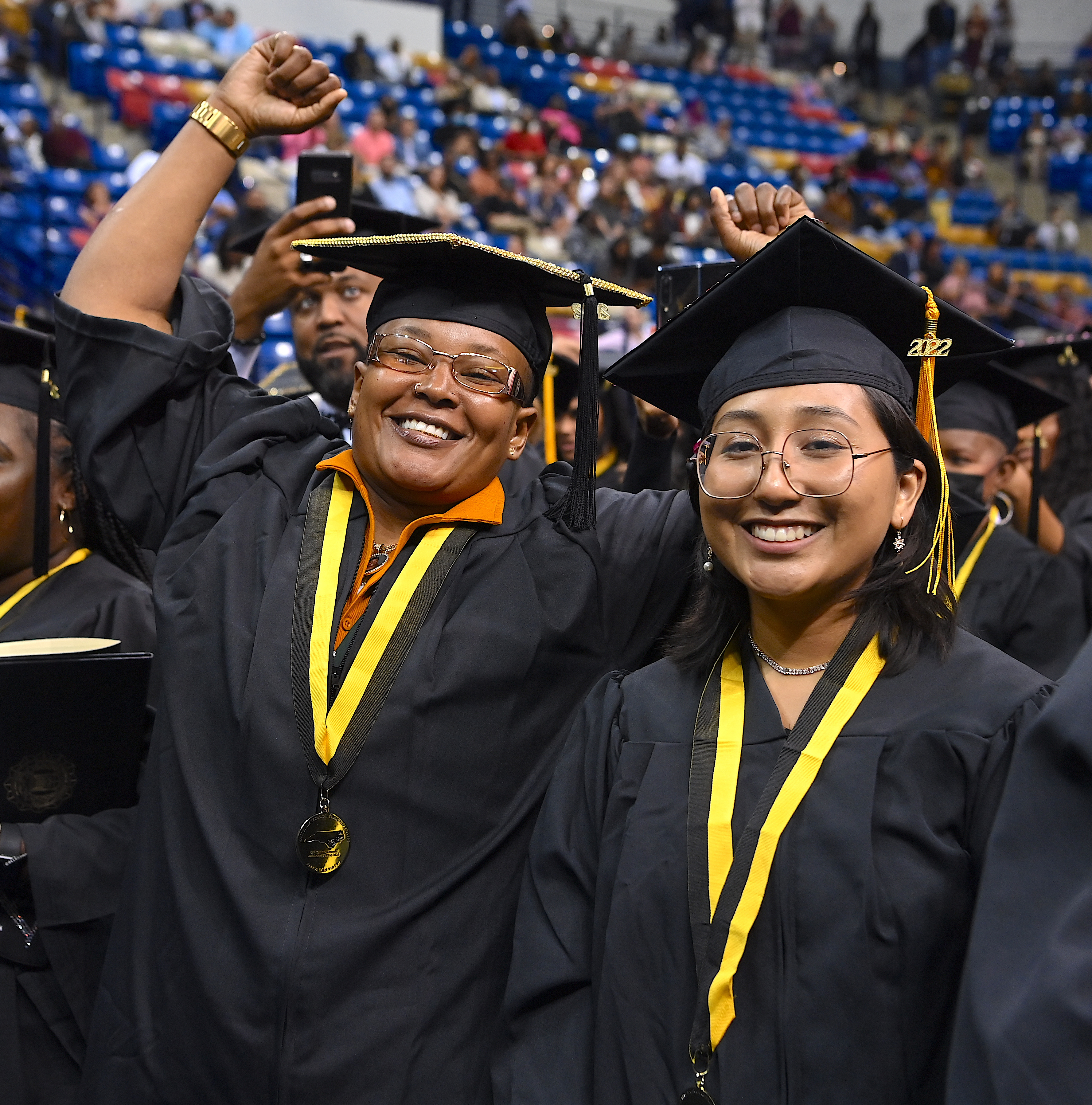 Two graduates smile at the camera. One of them has her hands raised in celebration and triumph. 