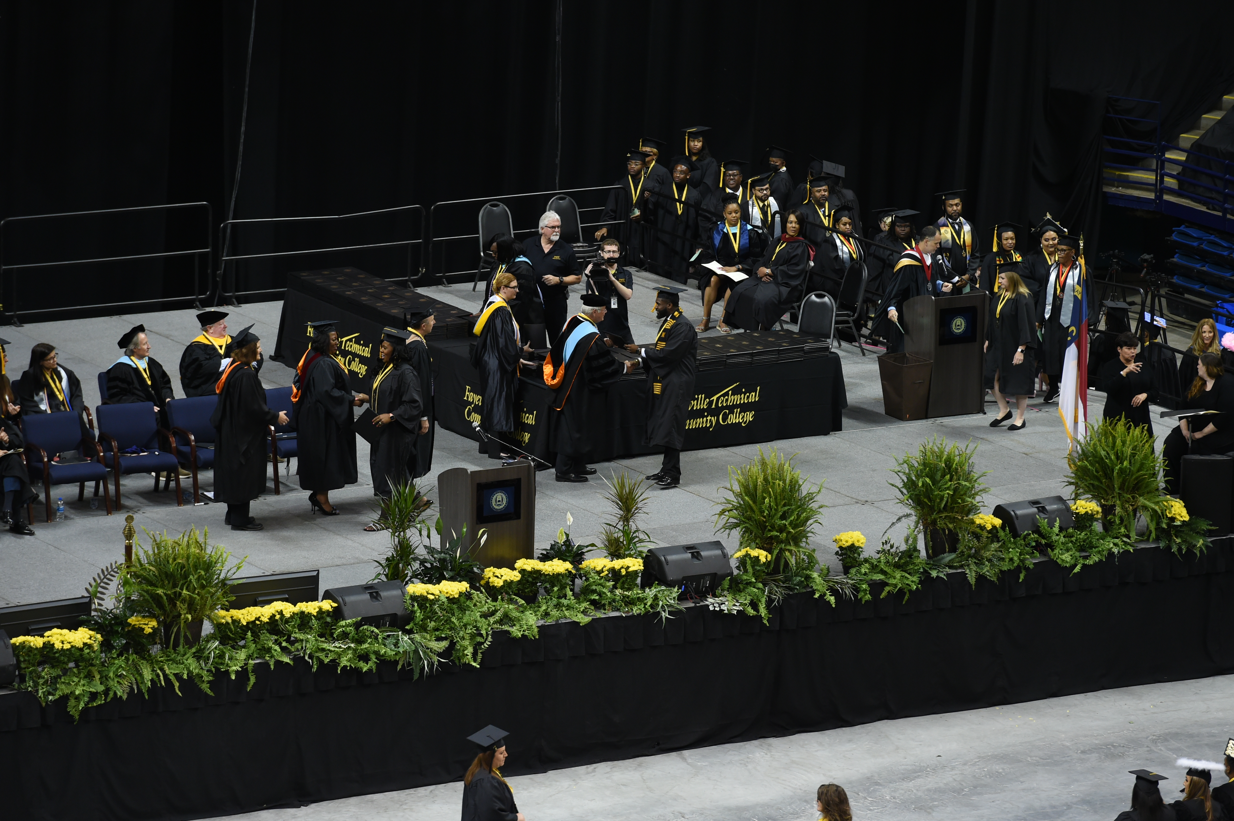 A wide photo of the stage as graduates come across to accept their degree portfolios and shake hands with faculty and staff.