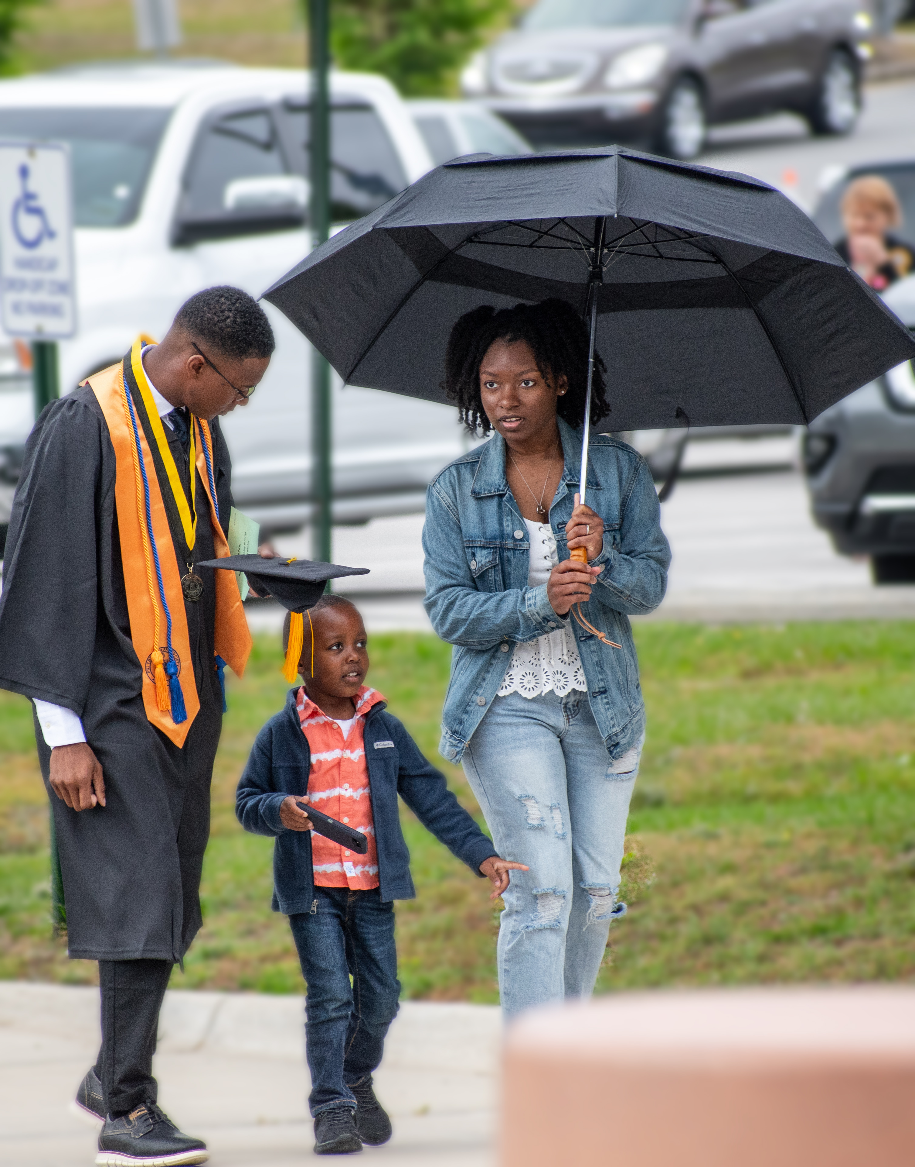 A graduate walks with his family. The graduate looks down at the young boy while the woman holds an umbrella over herself and the child.