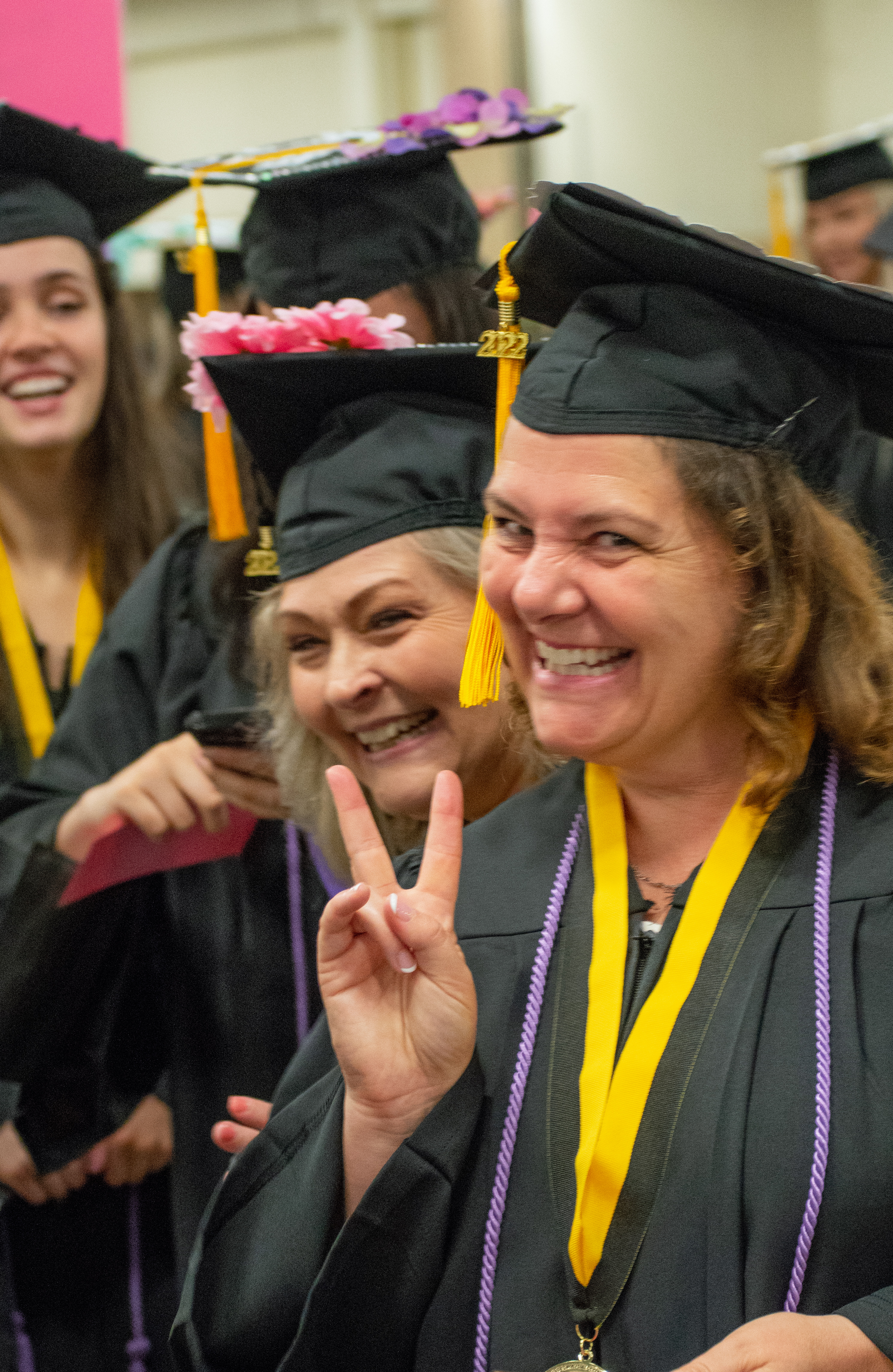 Two graduates smile at the camera. The graduate in the foreground is forming a peace sign with her fingers.