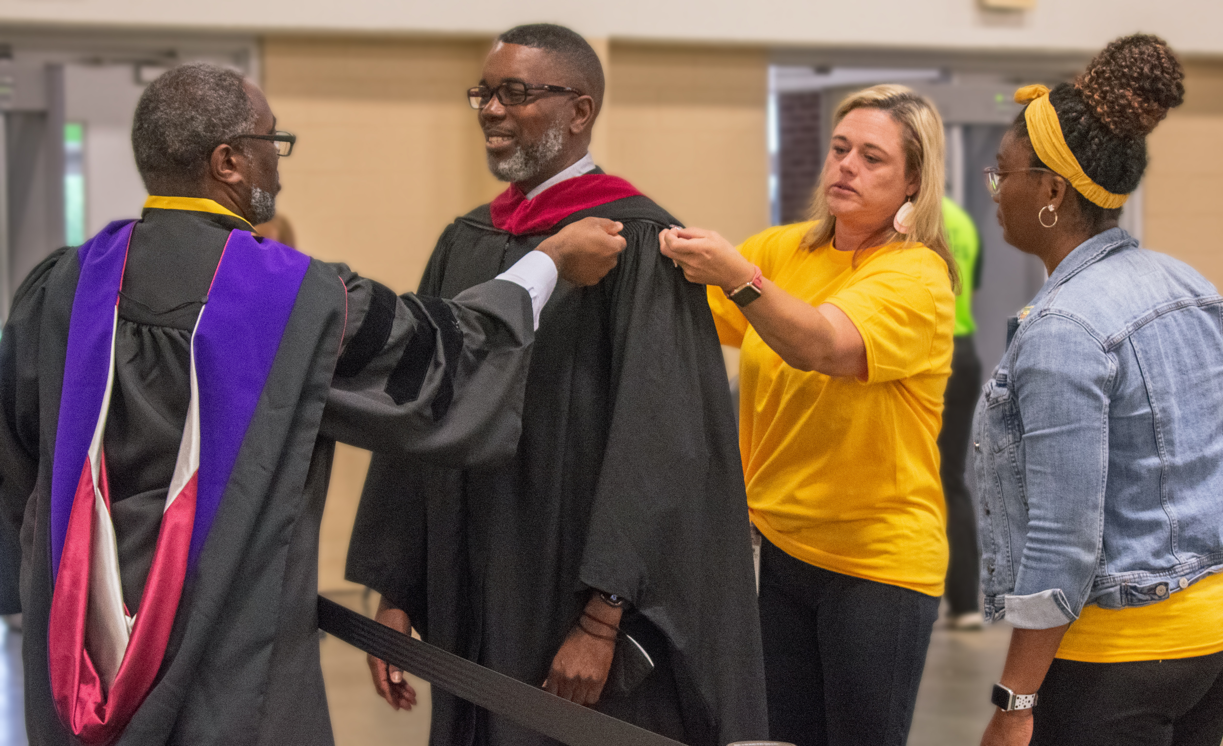 A man and woman help a man in graduation regalia adjust his hood as another woman looks on.