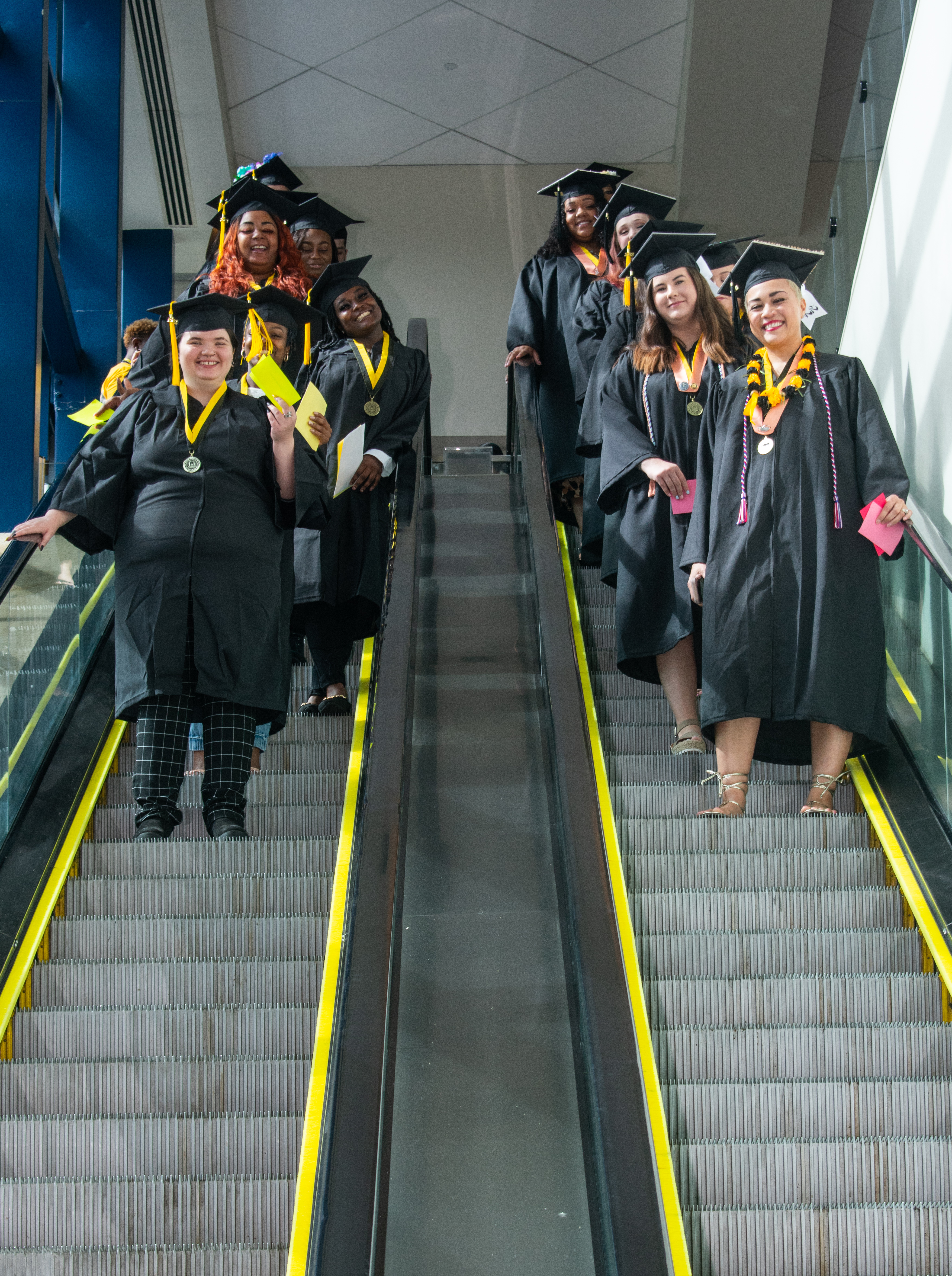 Graduates smile at the camera as they come down two escalators before graduation.