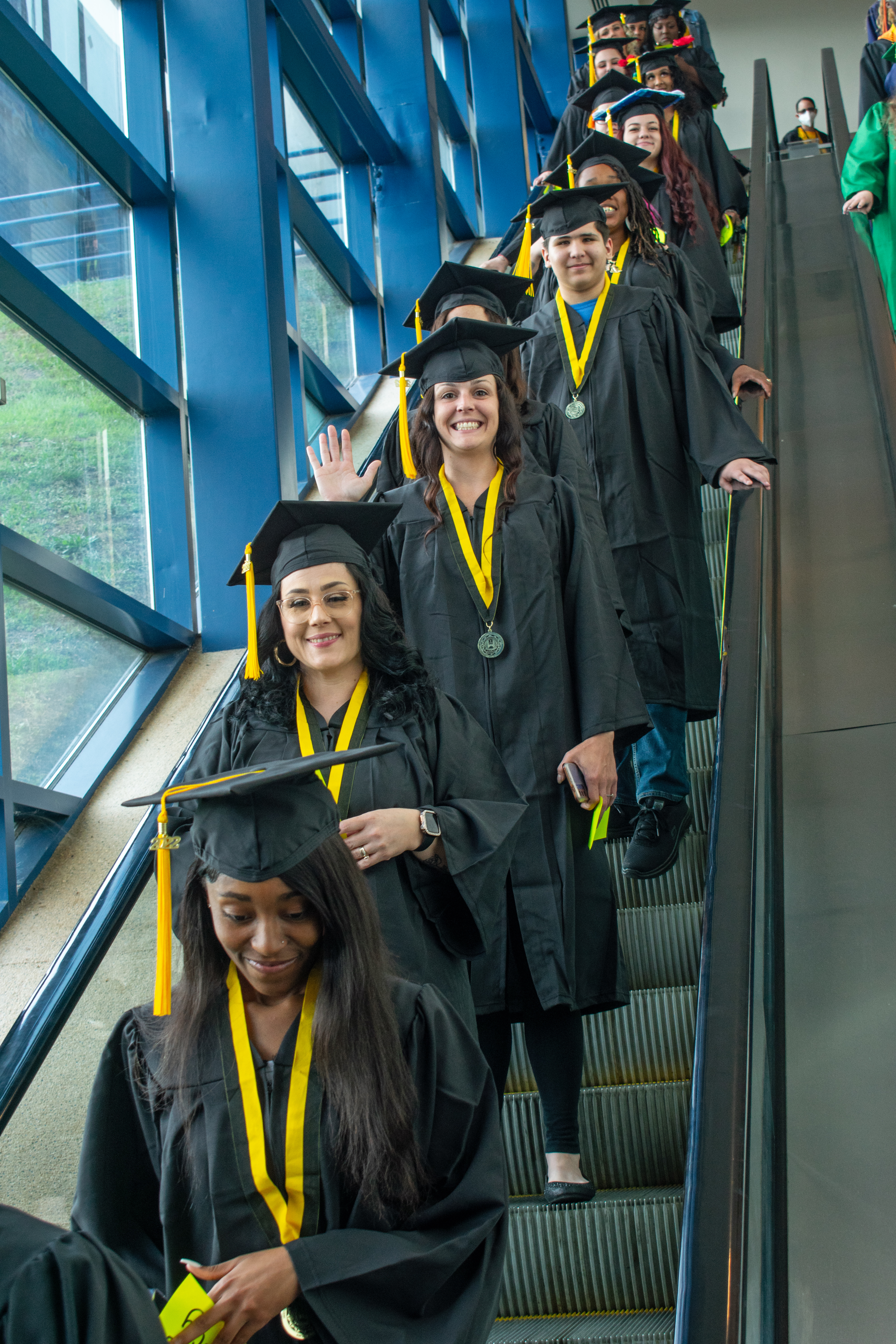 A line of graduates smile at the camera as they come down an escalator.