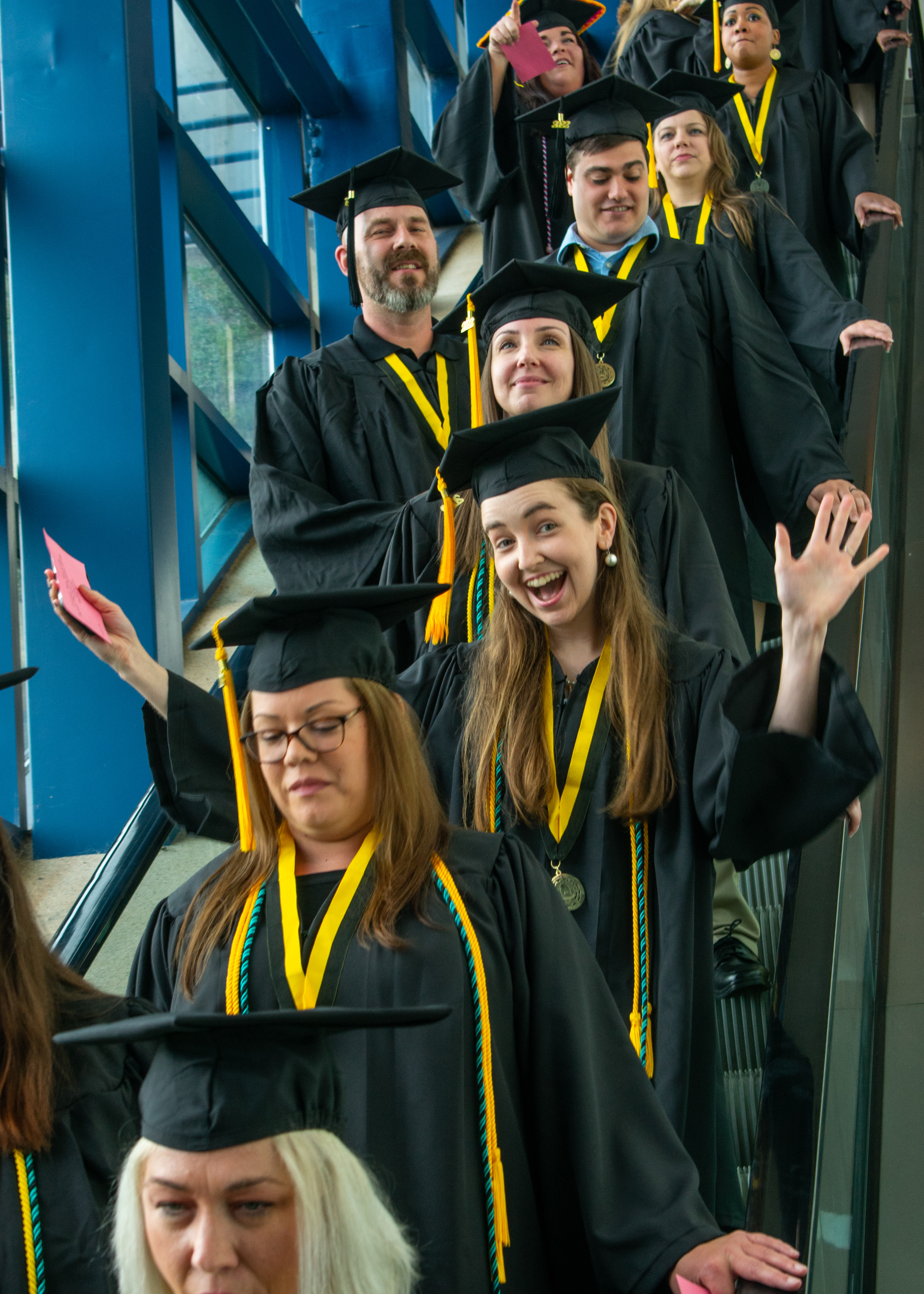 A graduate standing in a line of graduates coming down an escalator holds her arms in the air in a display of enthusiasm.