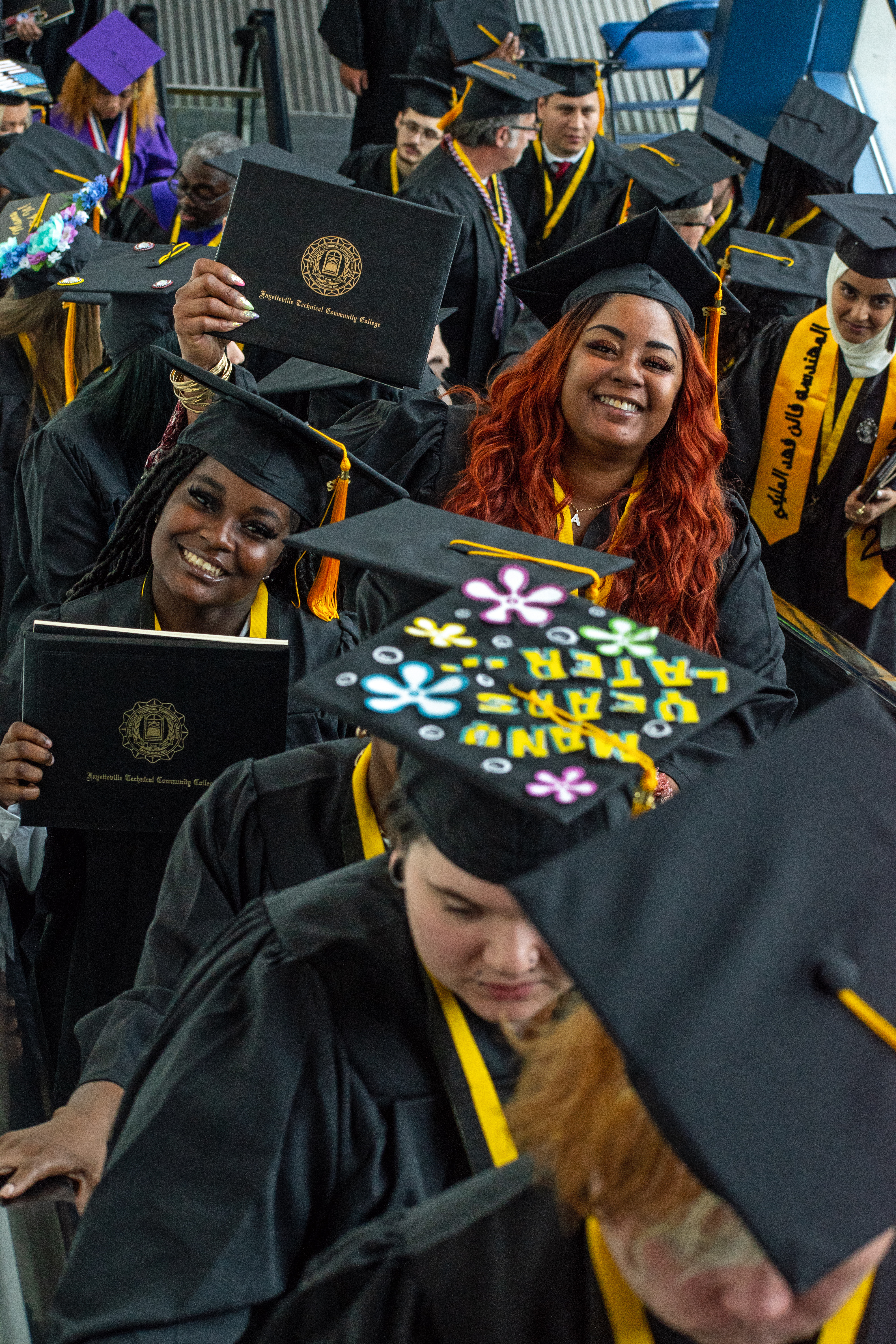 Two graduates in the background of a group of graduates hold up their degree portfolios and smile at the camera.
