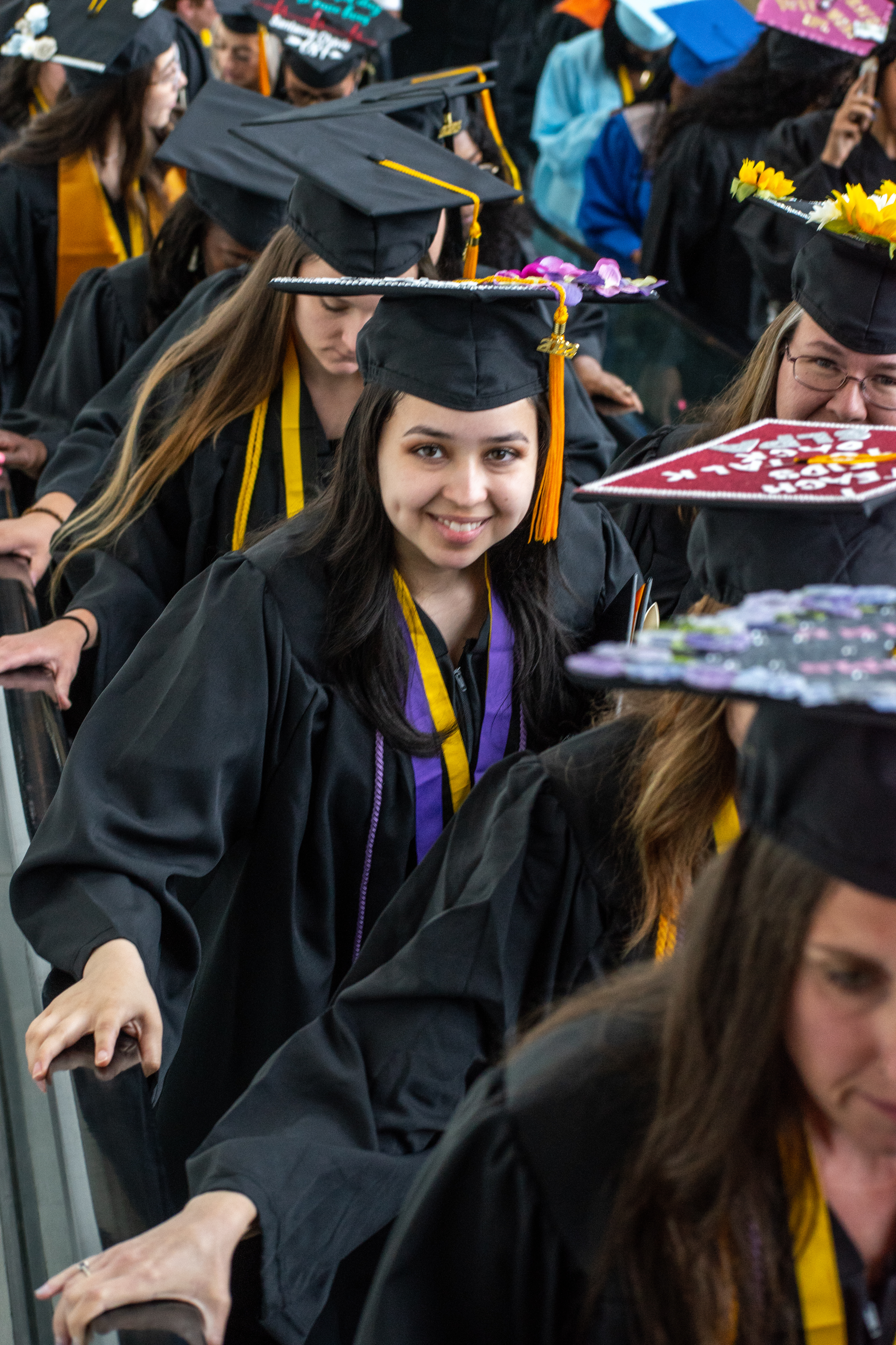 A graduate looks up and smiles at the camera as she goes up an escalator. She is surrounded by graduates.