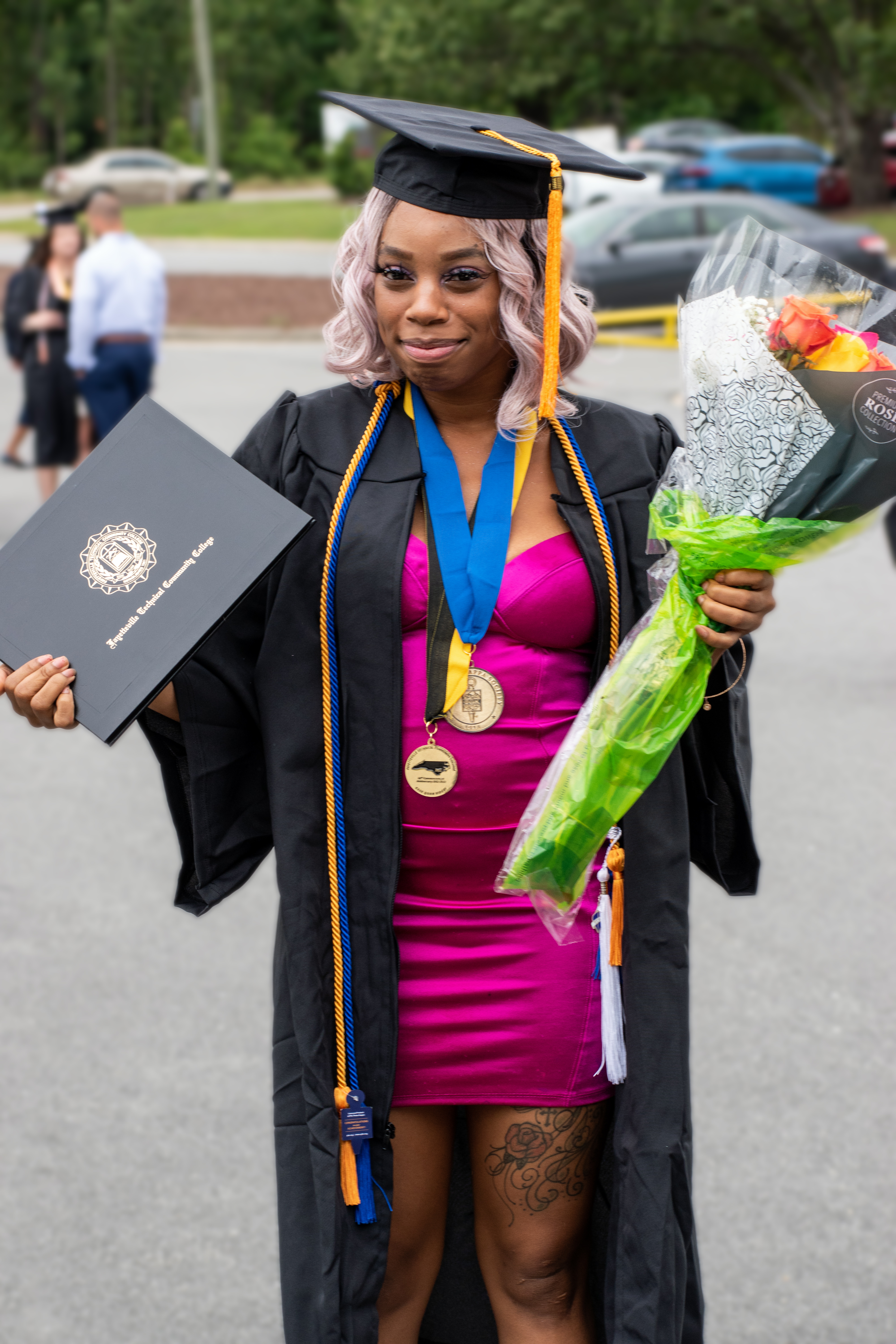 A graduate with pink hair has her gown open to reveal a pink dress. She is standing outside and is holding her degree portfolio and a bouquet of flowers.