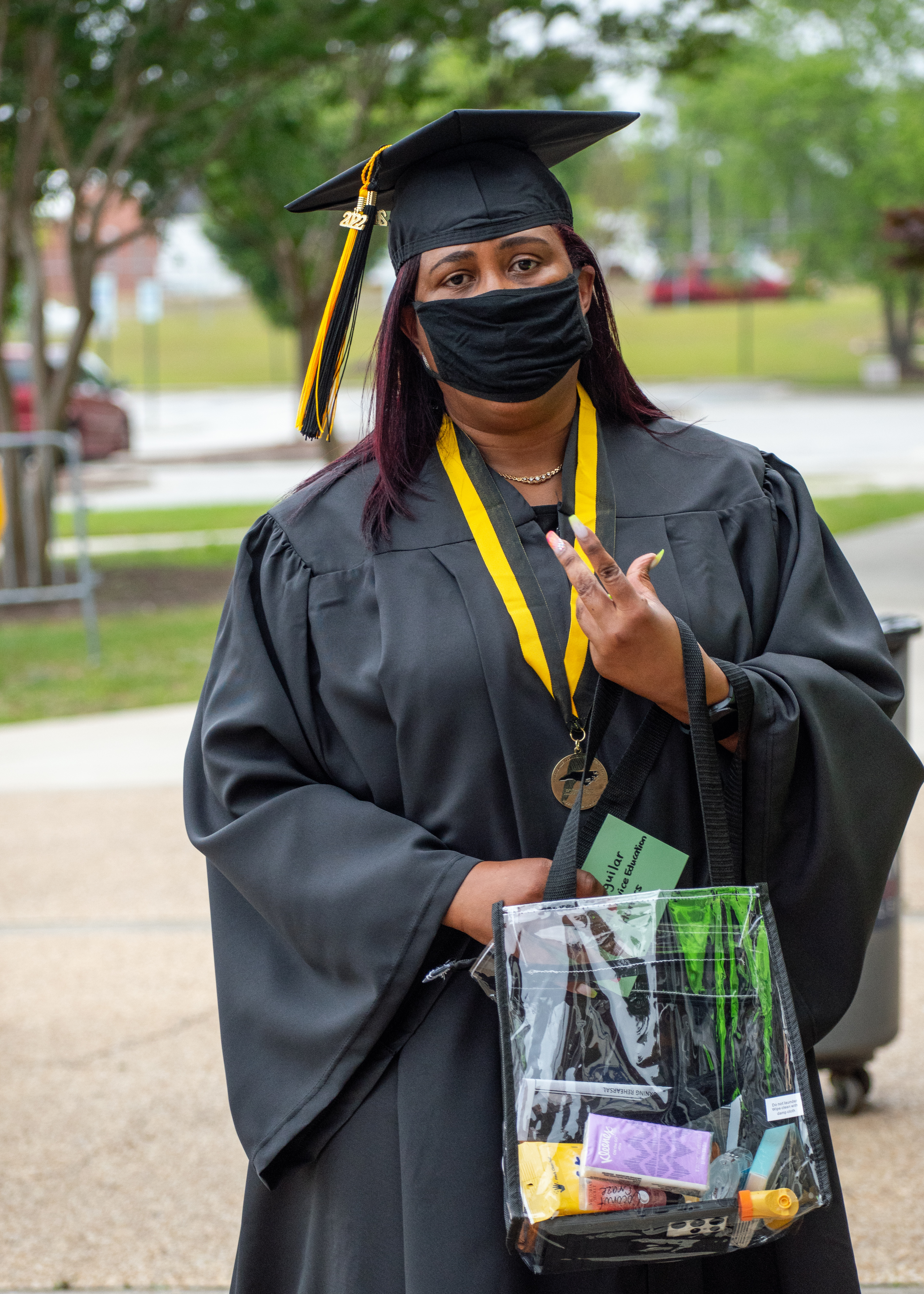 A graduate walks up the sidewalk. She is holding up two fingers on one hand.