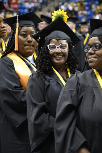 A graduate wearing white-framed glasses smiles at the crowd. She is surrounded by other graduates.