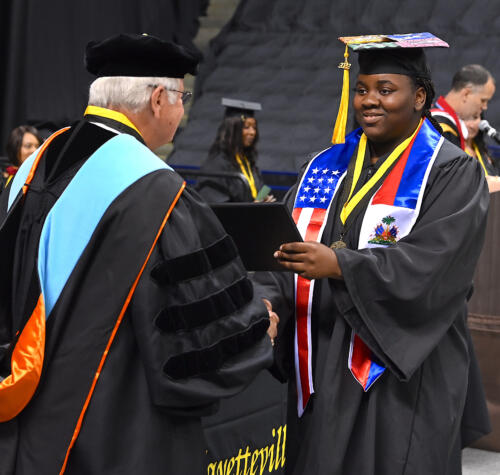 A graduate smiles as he accepts his degree portfolio from Dr. Keen. The graduate is wearing a large red, white and blue stole, honoring his military service.