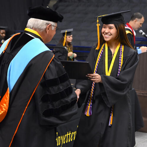 A graduate smiles as she accepts her degree portfolio from Dr. Keen. She has a gold cord and a medal on a black and gold ribbon hanging around her neck.