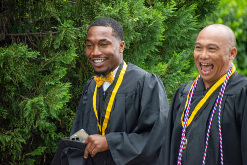 Two graduates without their caps share a laugh as they walk outside. Green trees are visible in the background.