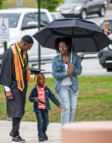 A graduate walks with his family. The graduate looks down at the young boy while the woman holds an umbrella over herself and the child.
