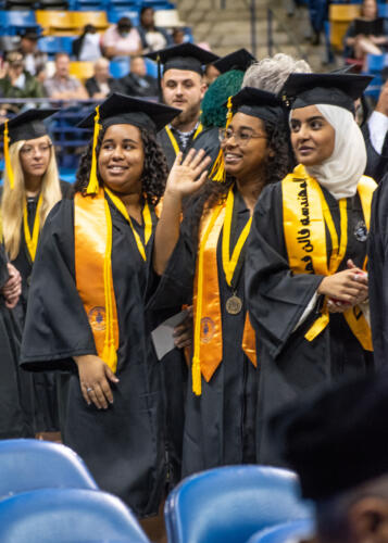 Three women, all wearing gold stoles with their caps and gowns, look up into the crowd. The graduate in the middle is waving.