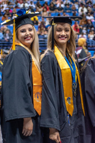 Two graduates with blond hair smile at the camera. They are both wearing gold stoles, and the graduate on the right has a medal on a blue ribbon around her neck.