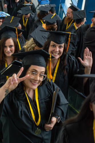 Three graduates, standing in a crowd of graduates, smile up at the camera. The graduate in the foreground is holding up two fingers to make a peace sign. The one on the right in the background is waving.