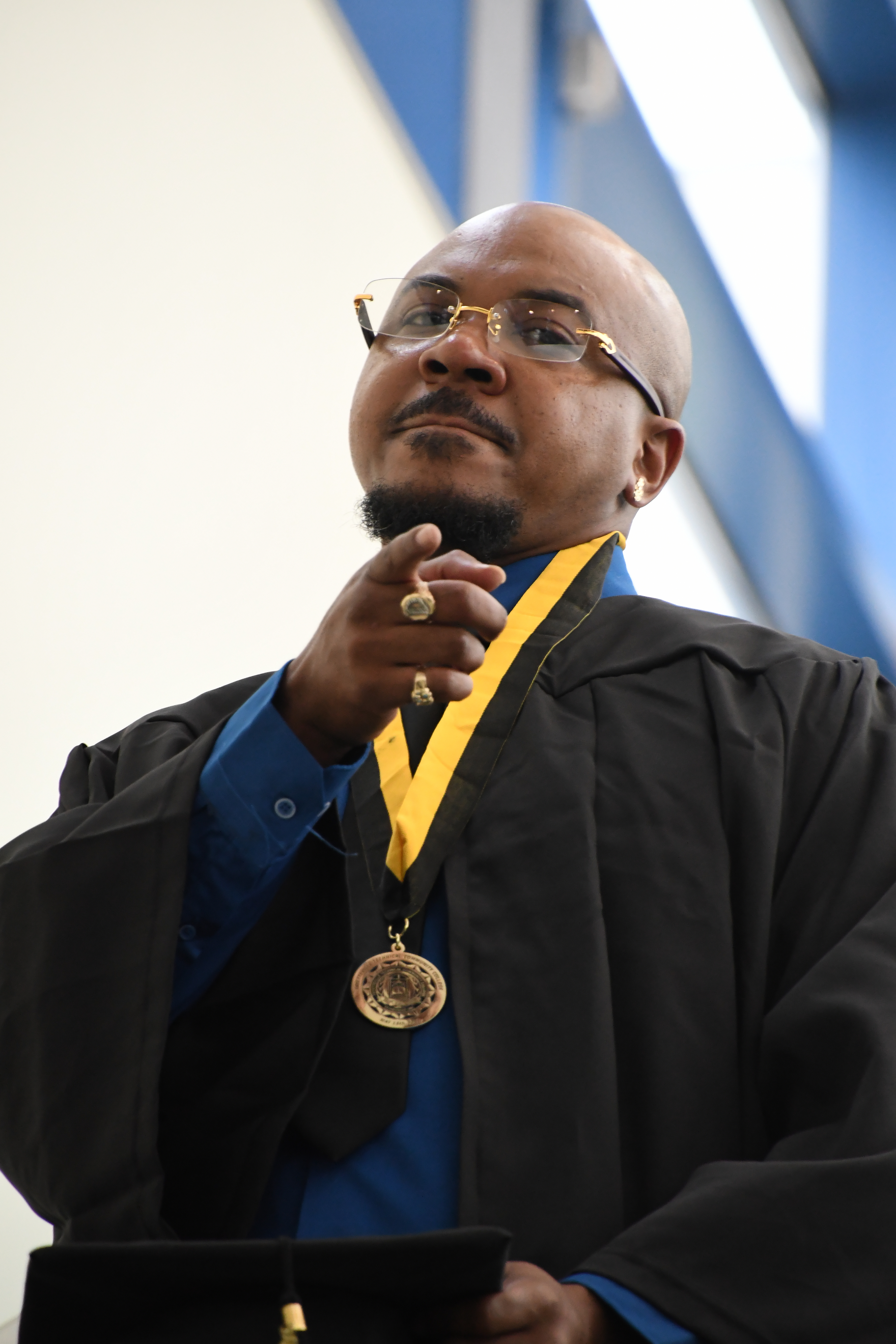A graduate without his cap looks down into and points at the camera as he comes down the escalator.