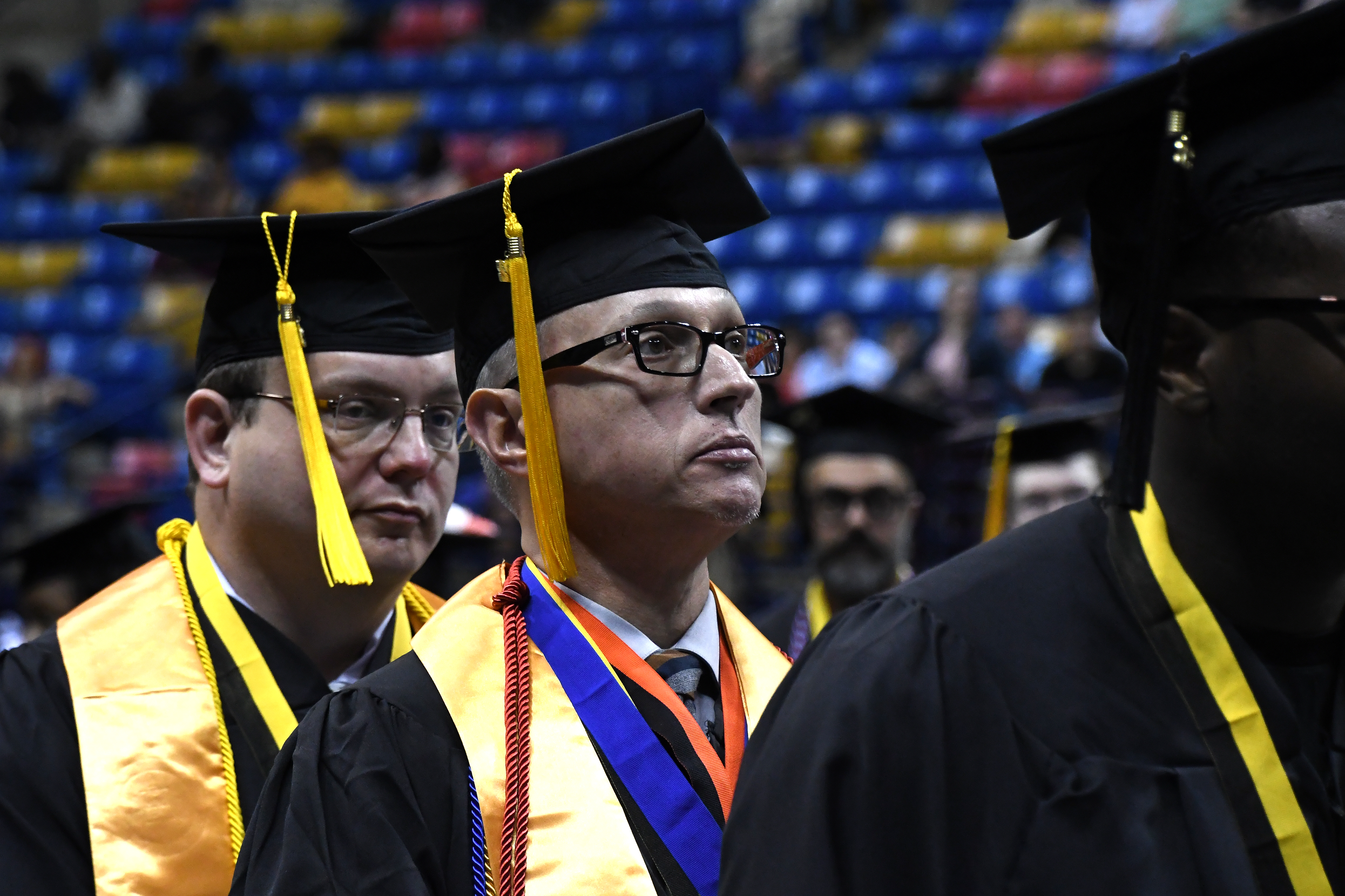 A close-up photo of a graduate wearing a gold stole, a red ribbon, a blue ribbon and a red cord around his neck.