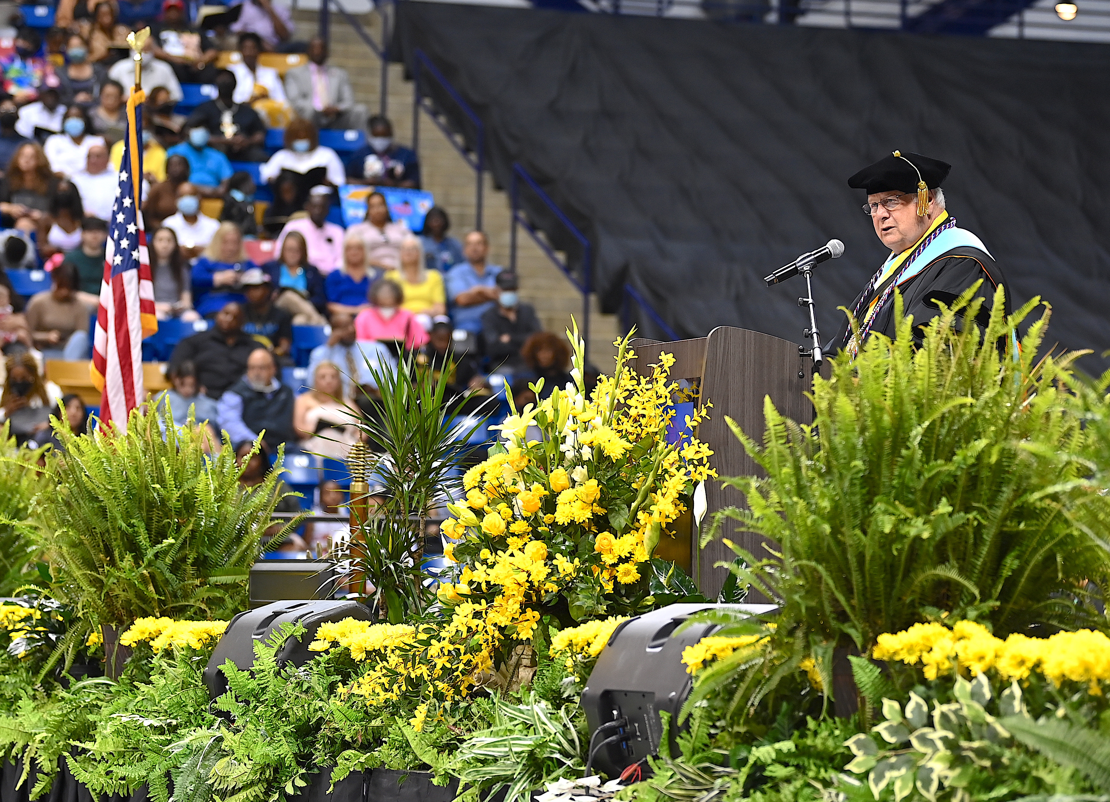 A wide photo of Dr. Keen speaking at the podium. Flowers and ferns at the front of the stage are visible in the foreground. Part of the audience is visible in the background. 