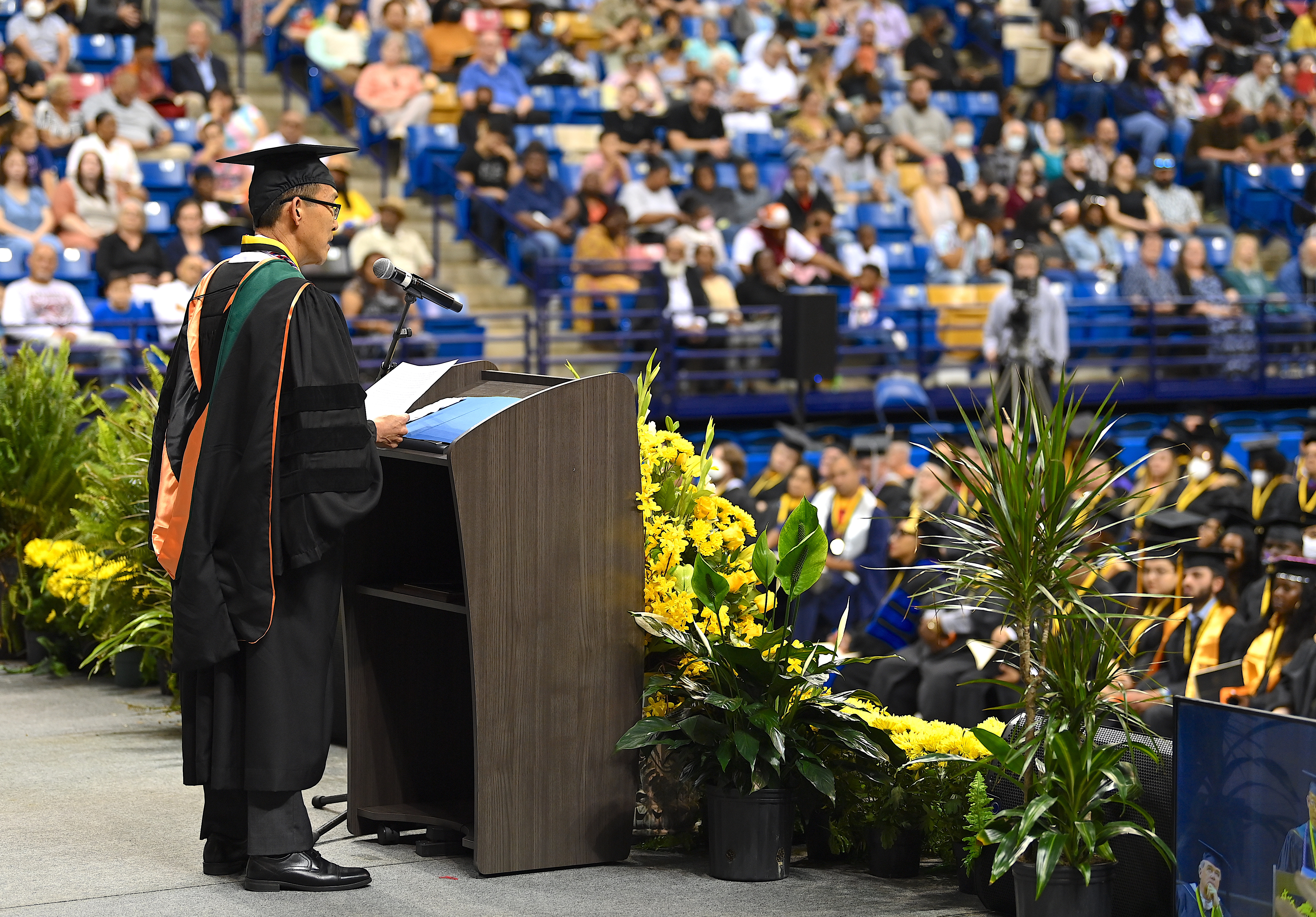 Dr. Choi, photographed from the side, speaks at the podium. Part of the graduates and the crowd is visible in the background.