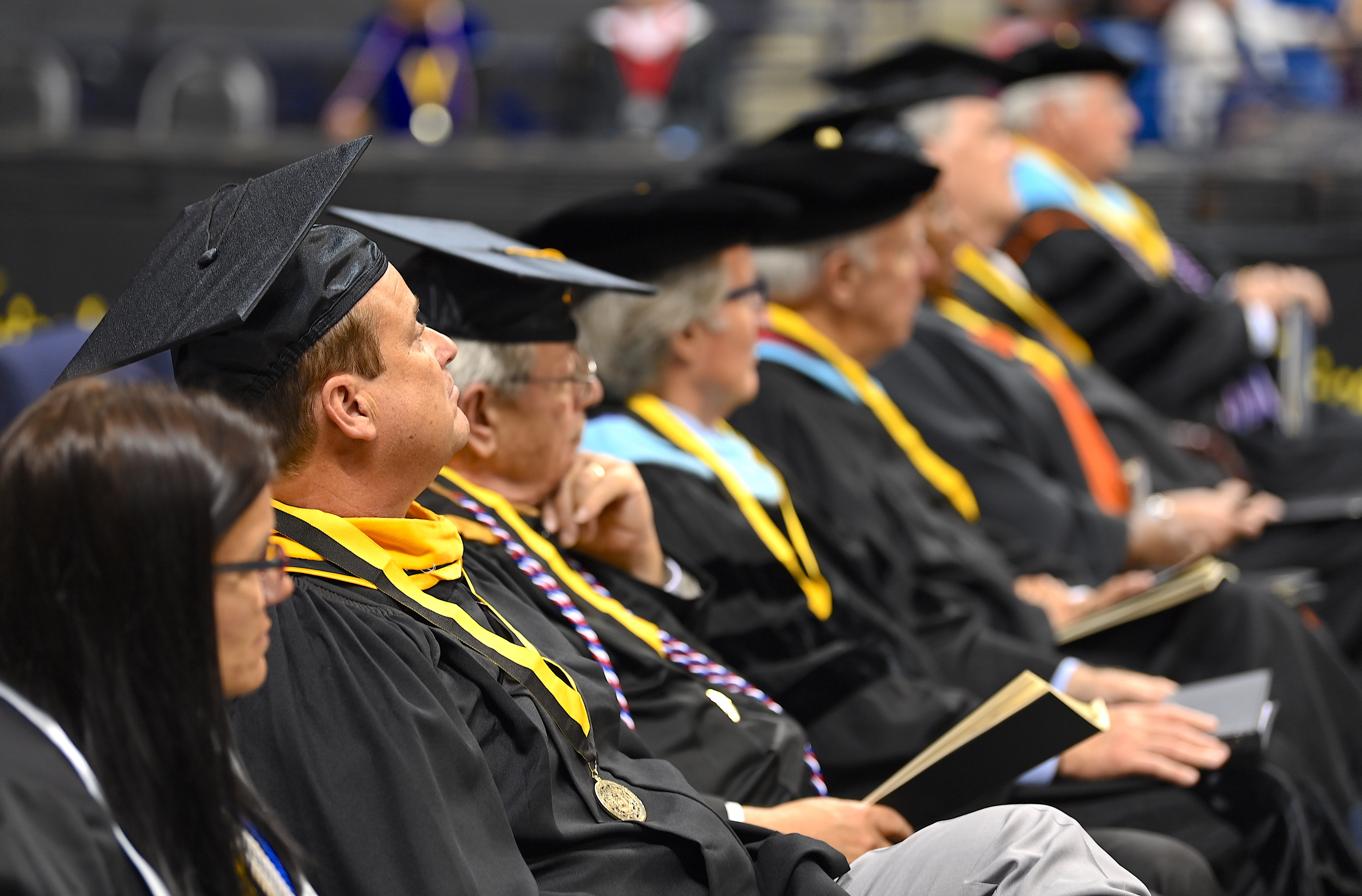 Faculty and staff, dressed in graduation regalia and seated on stage, look up towards the podium.